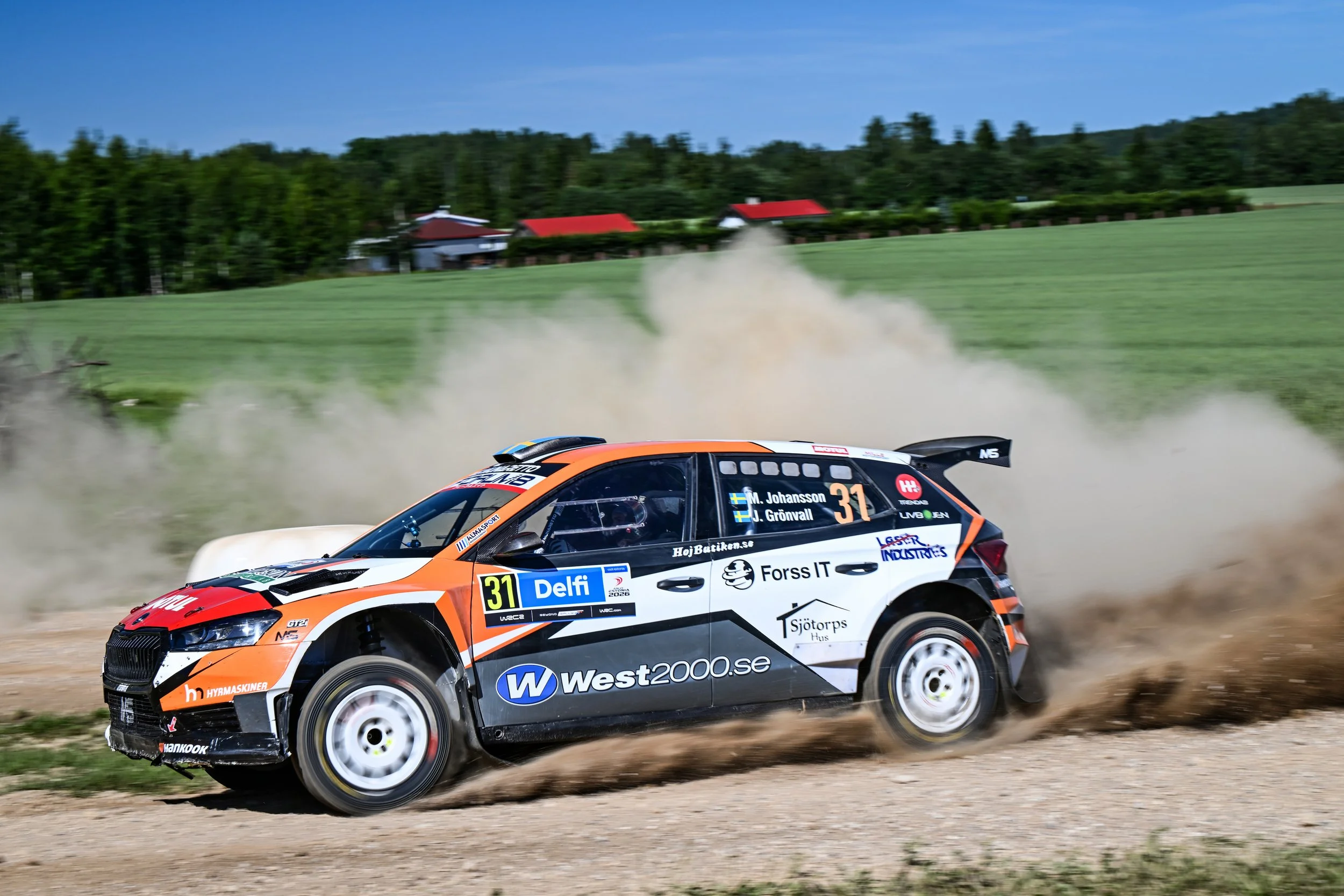 A rally car speeding on a gravel road with dust behind it, in a rural setting with green fields, trees, and houses with red roofs in the background.