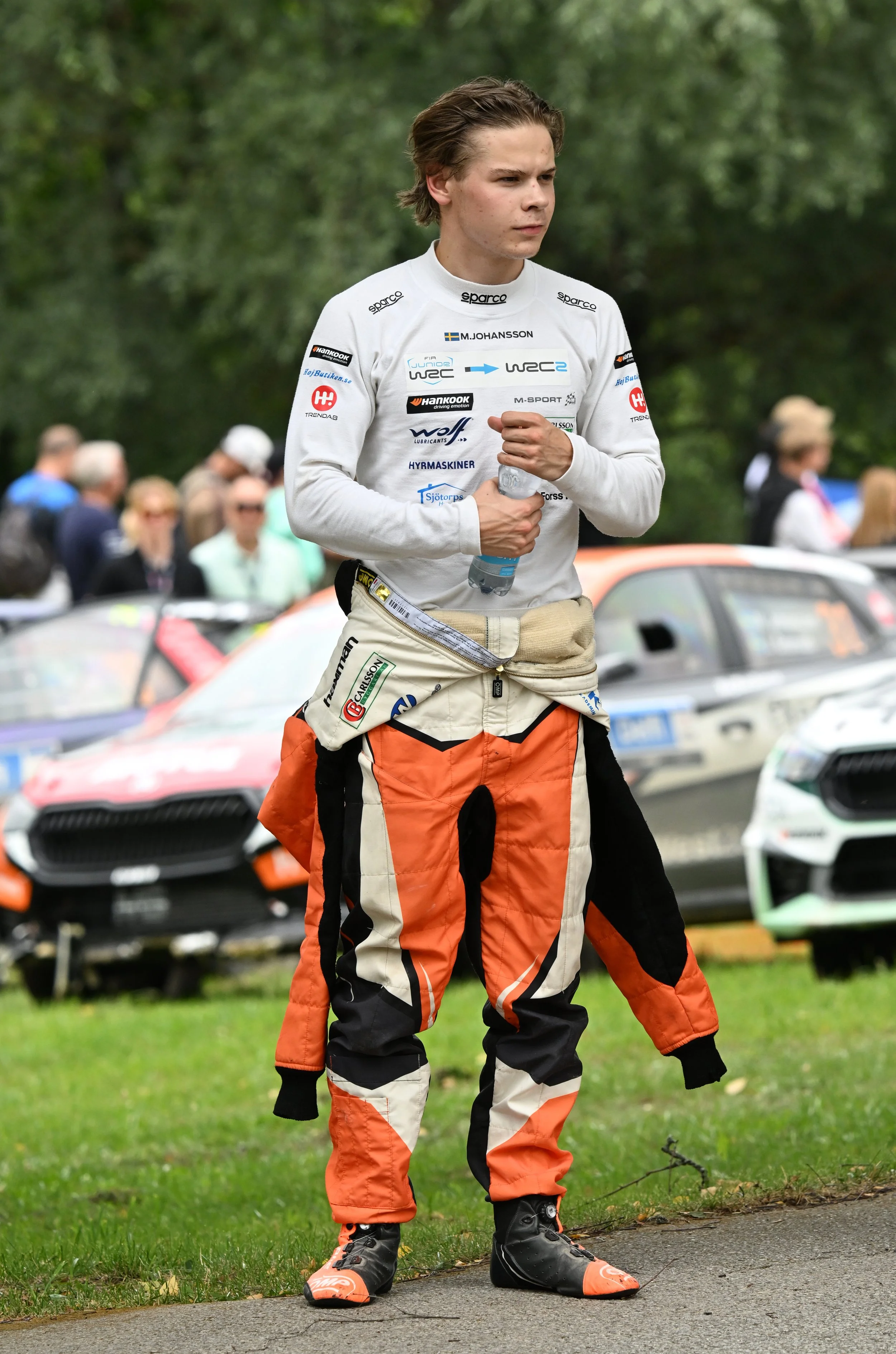A young man in a racing suit holding a water bottle, standing outdoors with rally cars and spectators in the background.