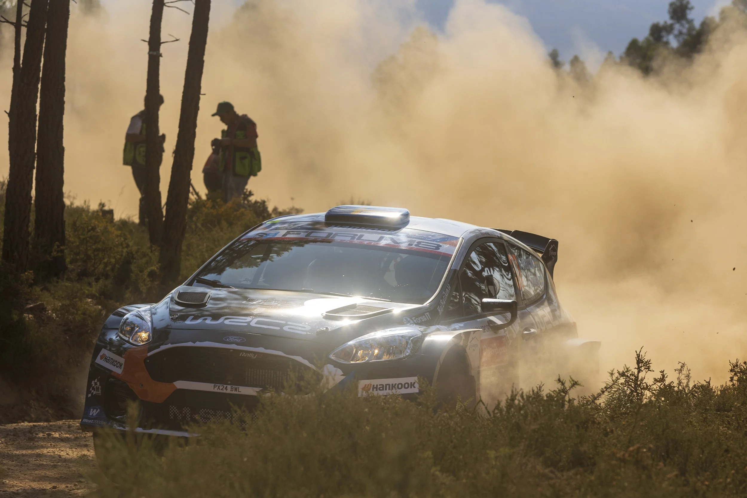 A rally car racing through a dirt road in a forest, kicking up dust in the air. Two people are seen standing among trees in the background, observing the race.