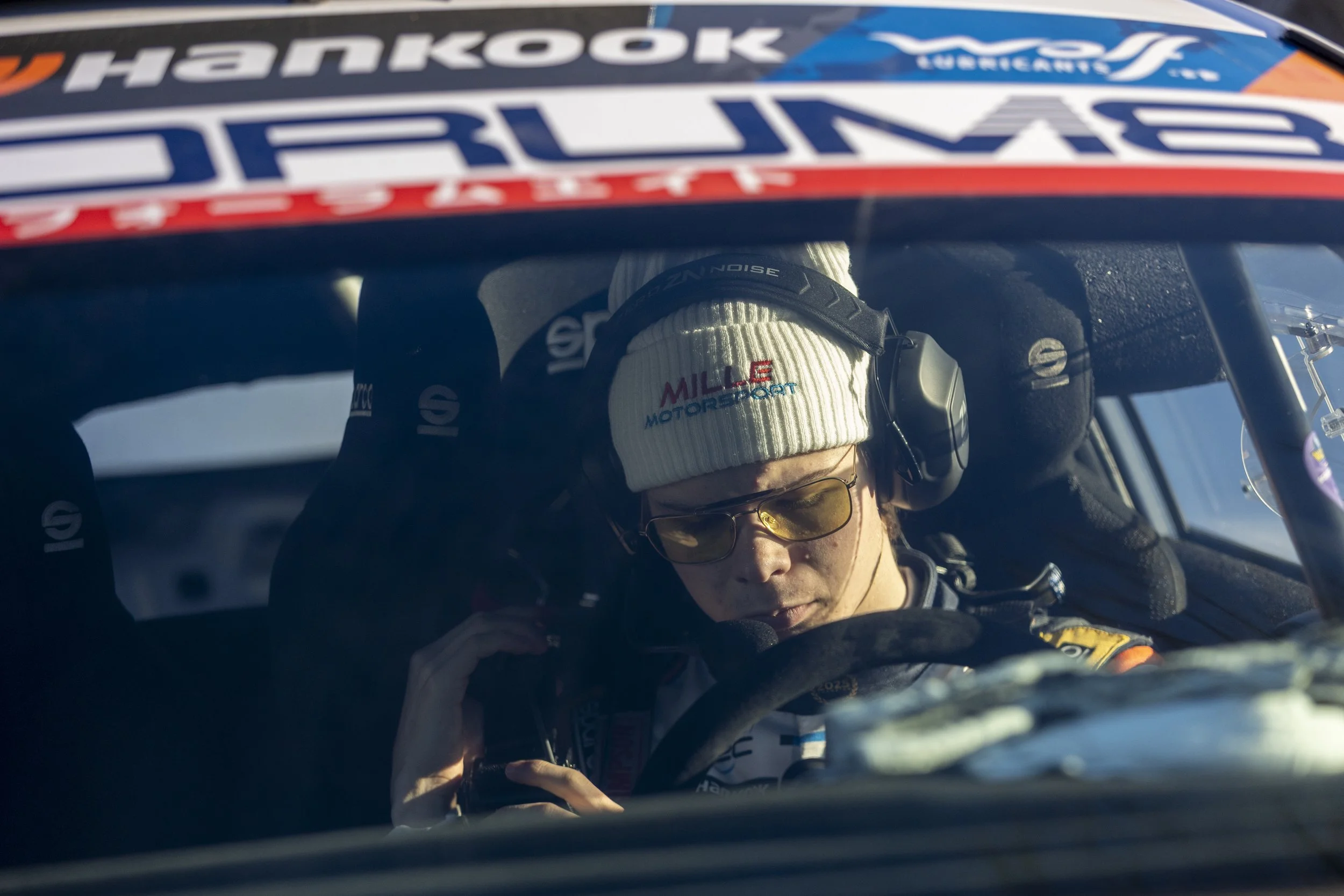 A person wearing a white beanie with red and blue text, glasses, and a racing suit, sitting in a race car cockpit. The scene is seen through the windshield with a logo and sponsor banners visible at the top of the image.