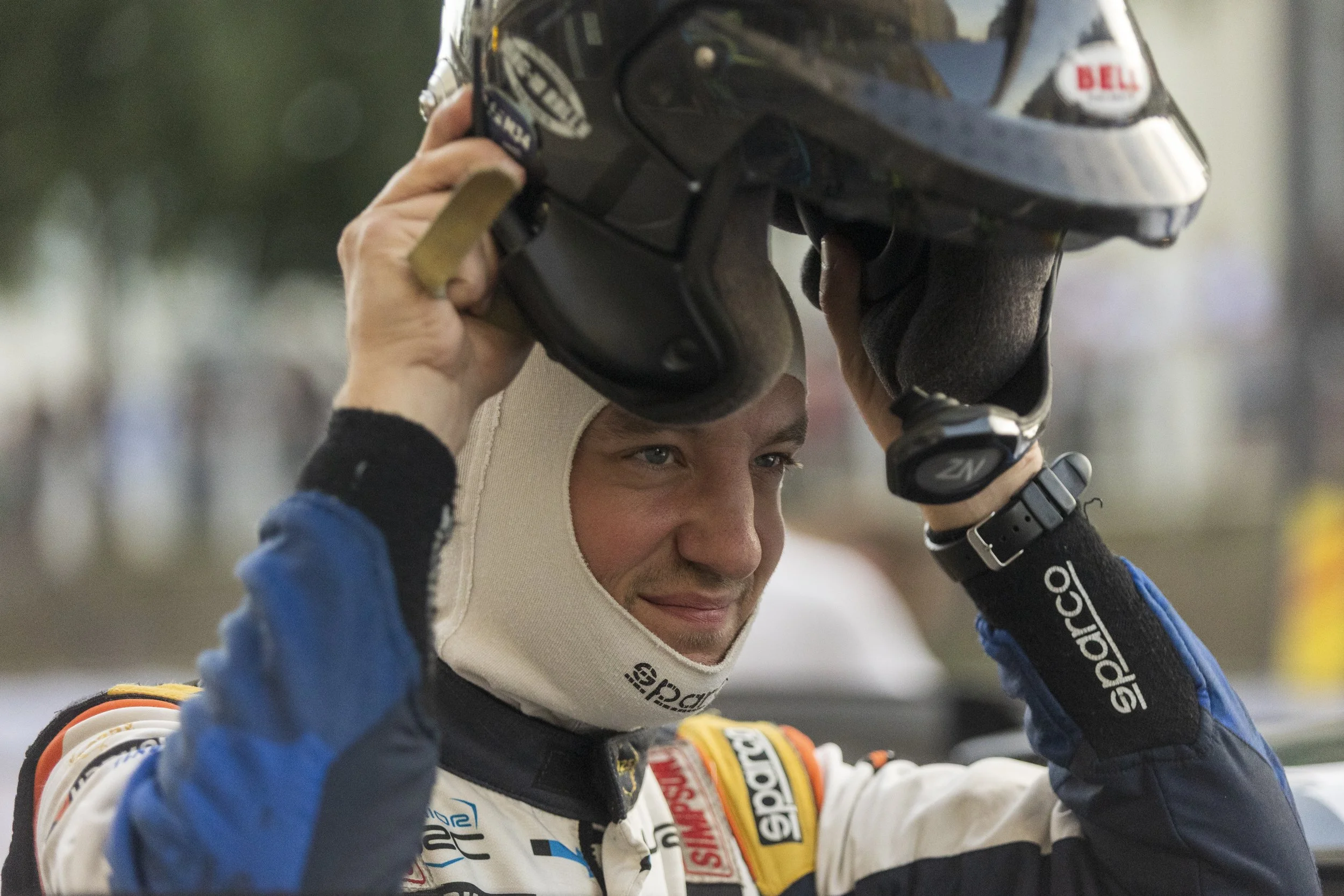 A race car driver is putting on a helmet, smiling, with racing suit and gloves, in a racing environment.