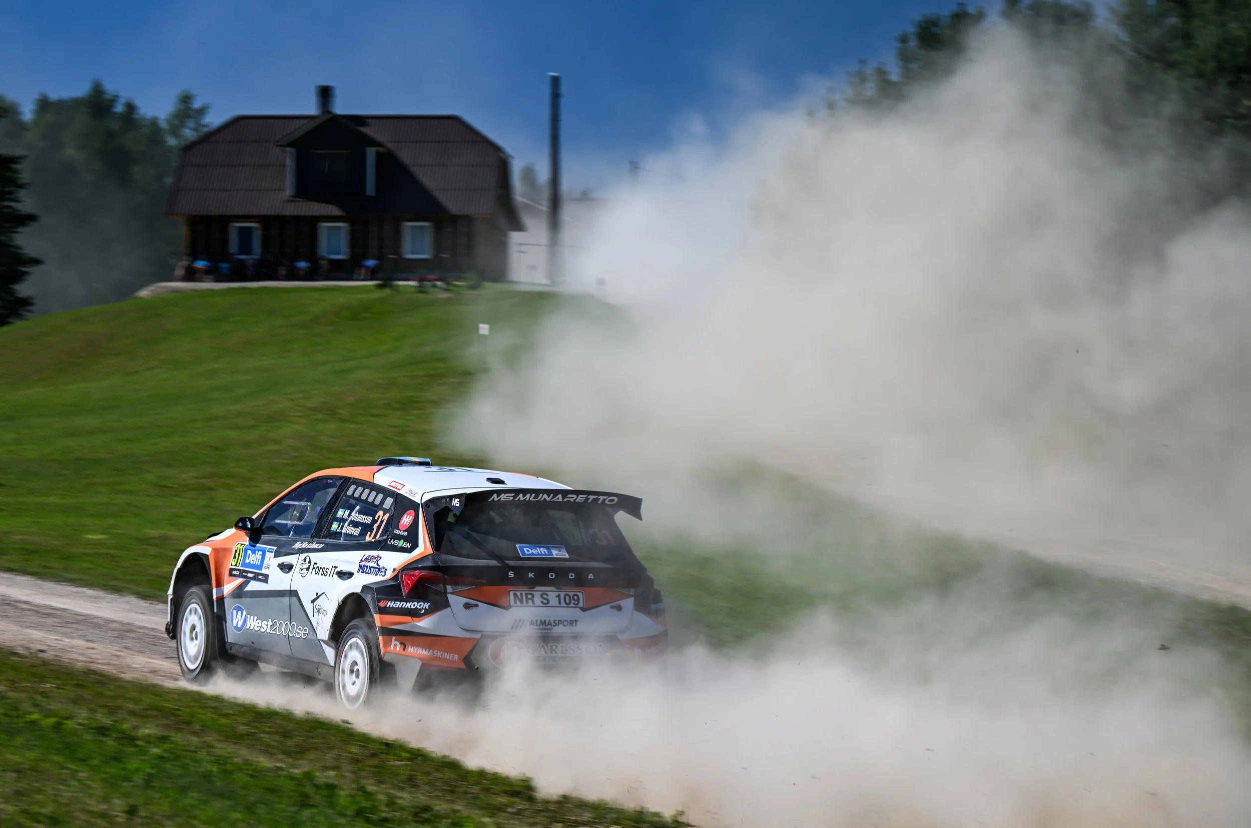 A rally car racing on a dirt road, kicking up a large cloud of dust, with a wooden house and trees in the background.