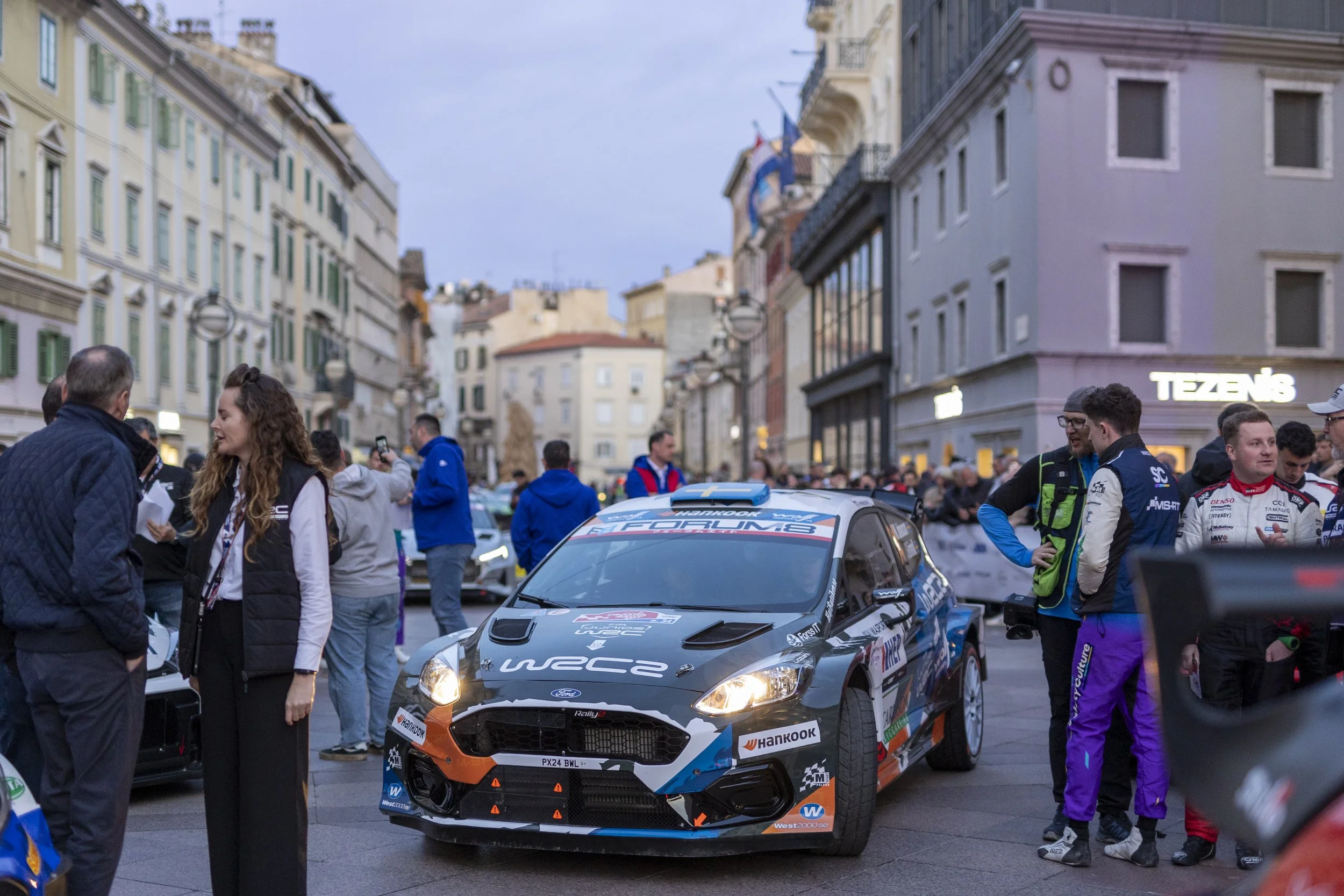 People gather around a rally car on a city street, with buildings in the background. Some individuals are taking photos and talking, likely at a motorsport event.