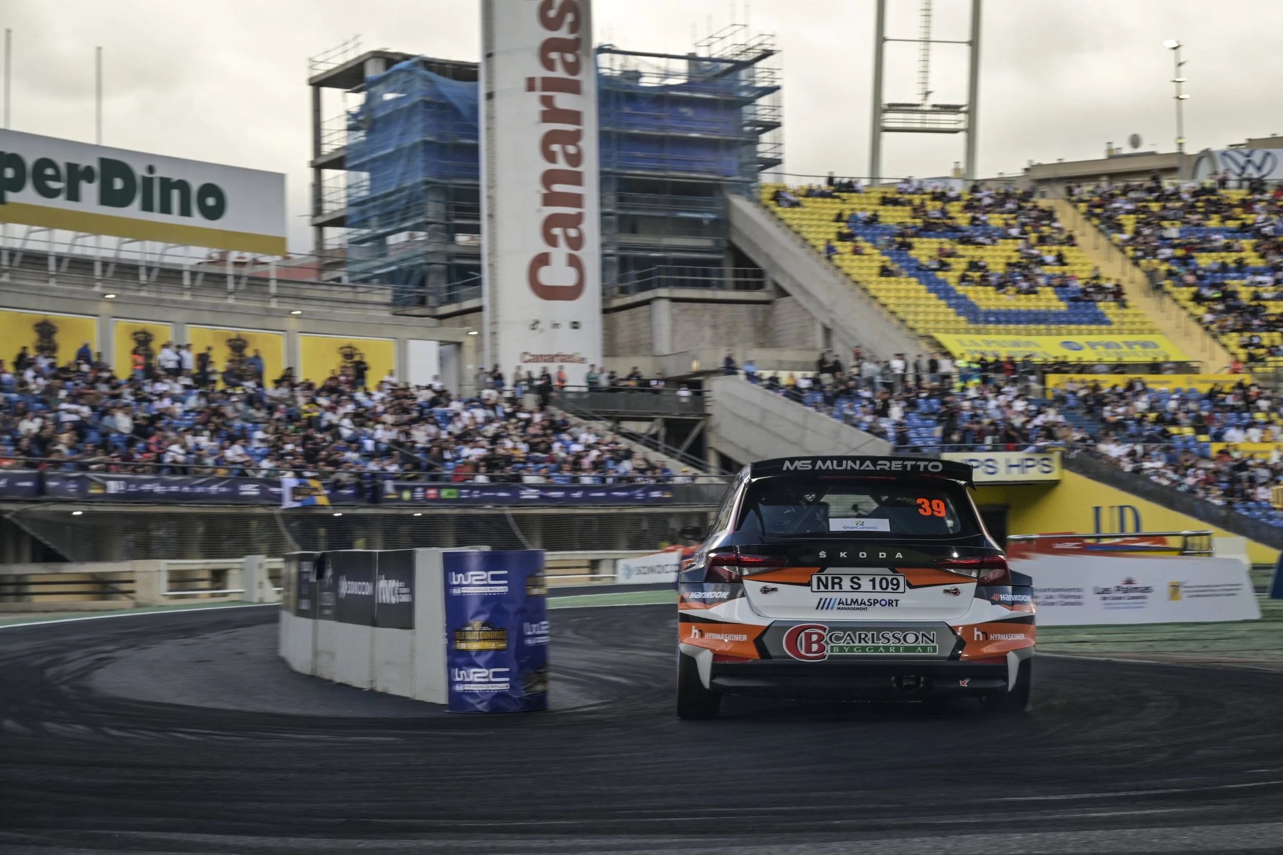 Race car on track with a crowd in the grandstands behind. The car has rally-style branding and sponsors, with the number 39 visible on the rear window.
