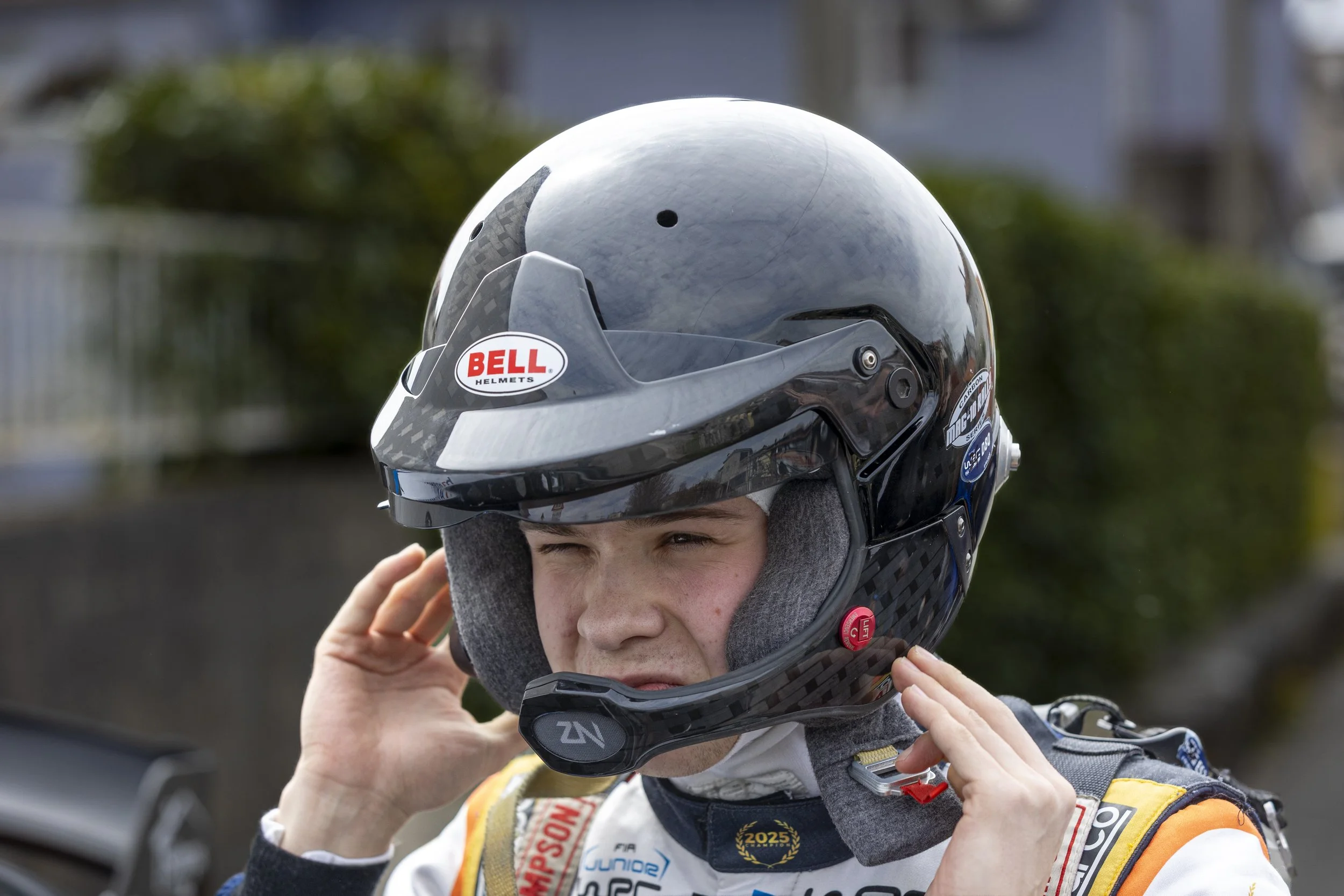 A young race car driver is adjusting their helmet before a race, wearing a white racing suit with sponsor logos.