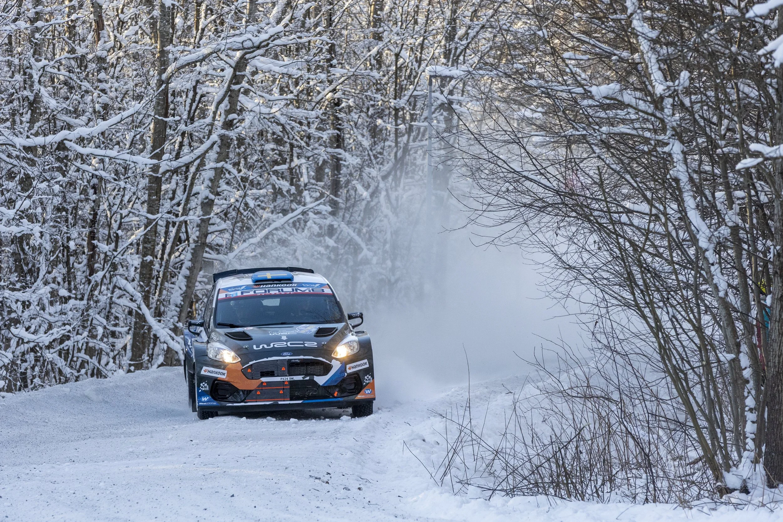 A rally car racing on a snowy forest trail with snow-covered trees and mist in the background.