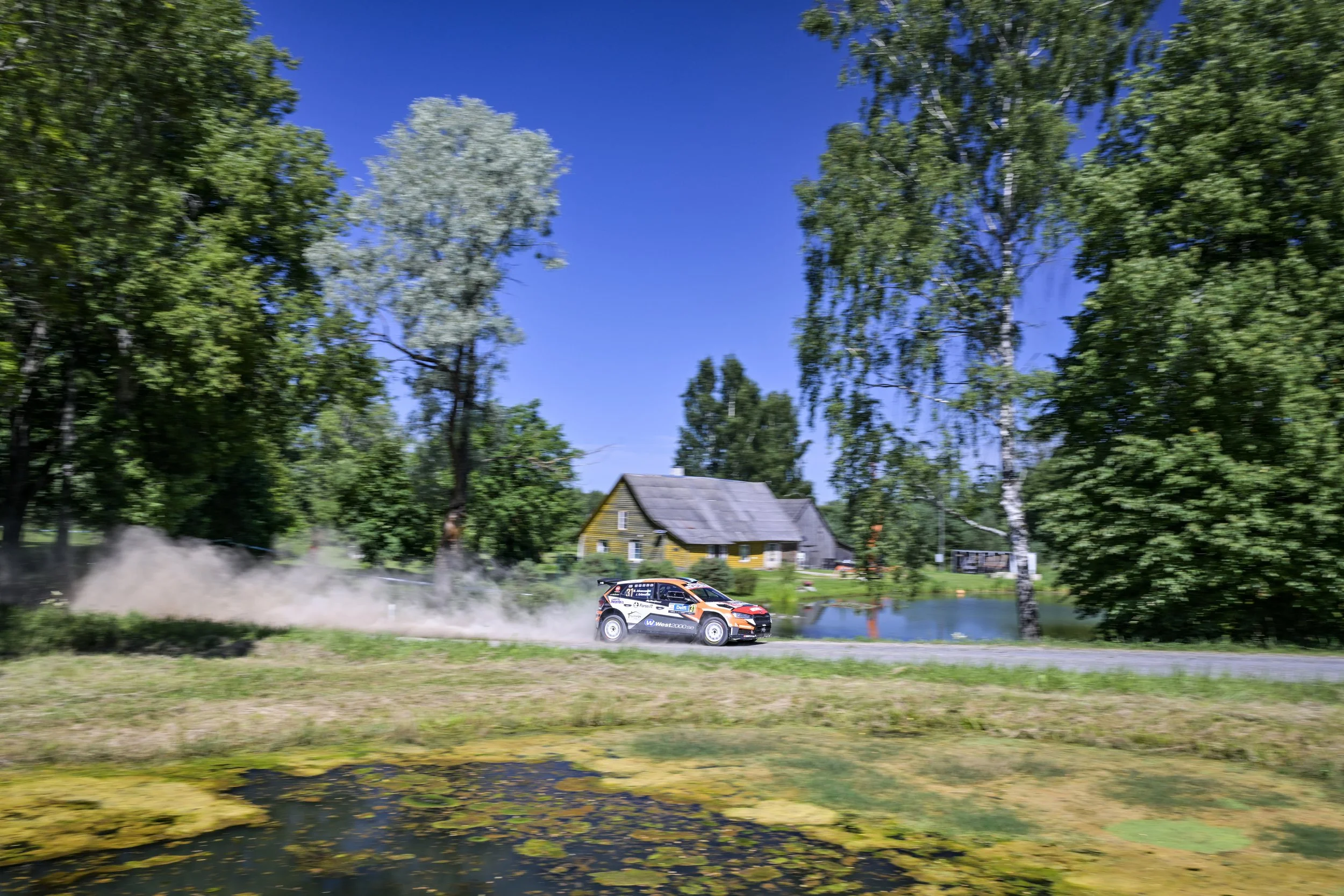 A rally car kicks up dust as it speeds along a rural dirt road, surrounded by green trees and houses by a pond under a bright blue sky.