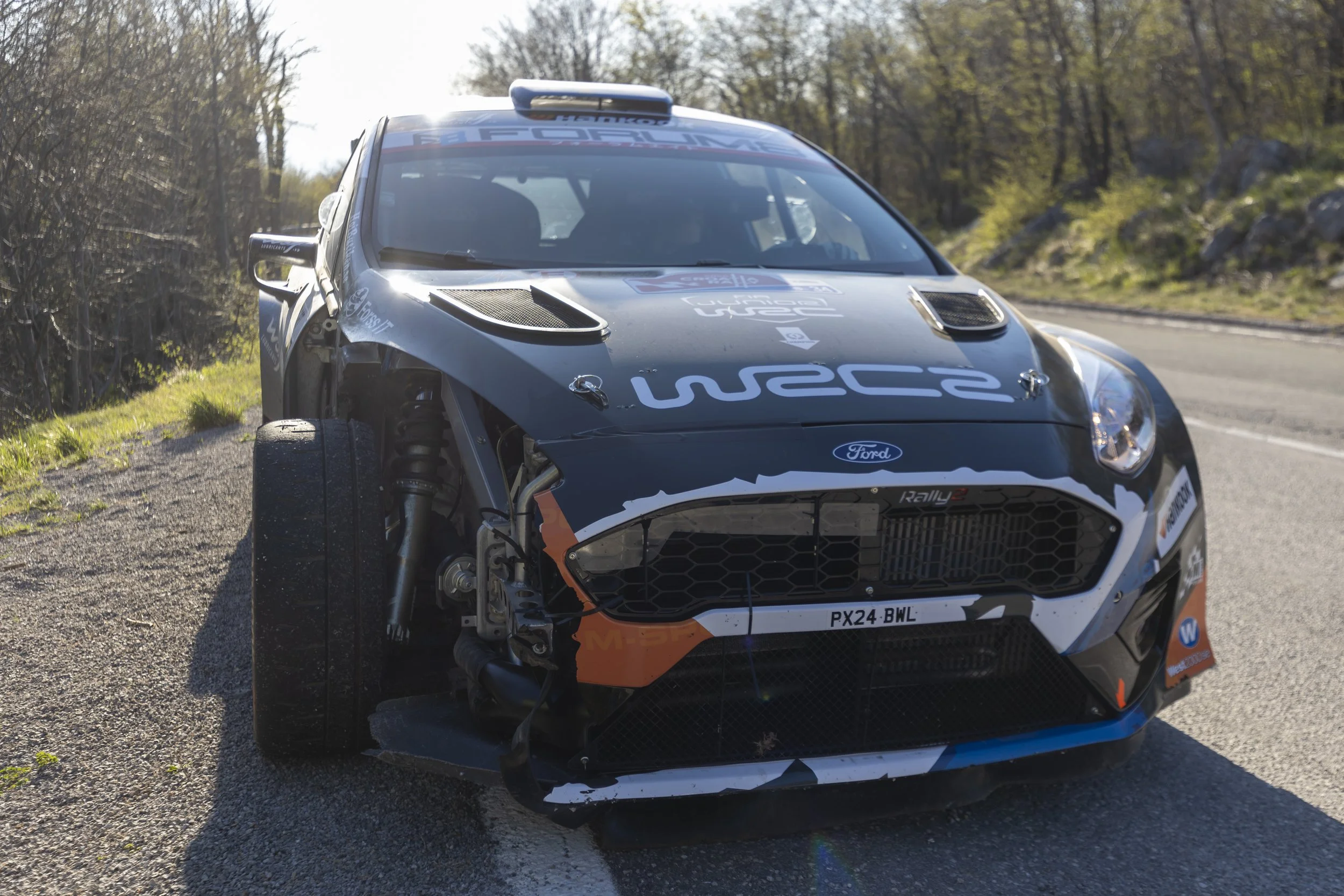 A partially damaged rally car on a gravel roadside with trees in the background.