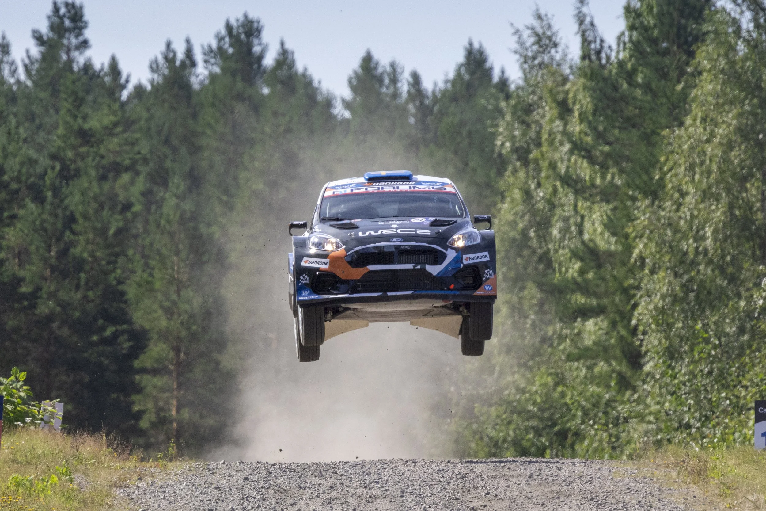 A rally car airborne above a dirt road in a forested area during a race.