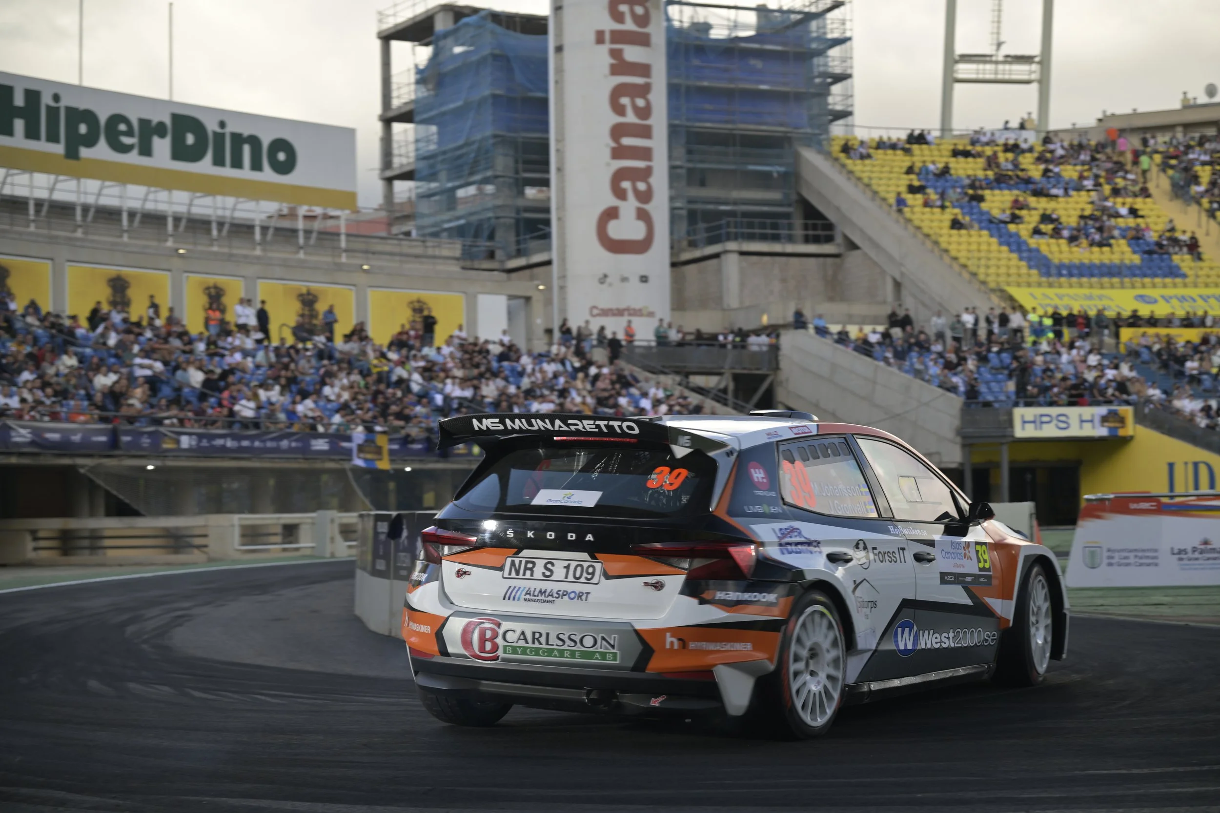 Race car on a racing track with a large crowd of spectators in the grandstands and advertising banners in the background.