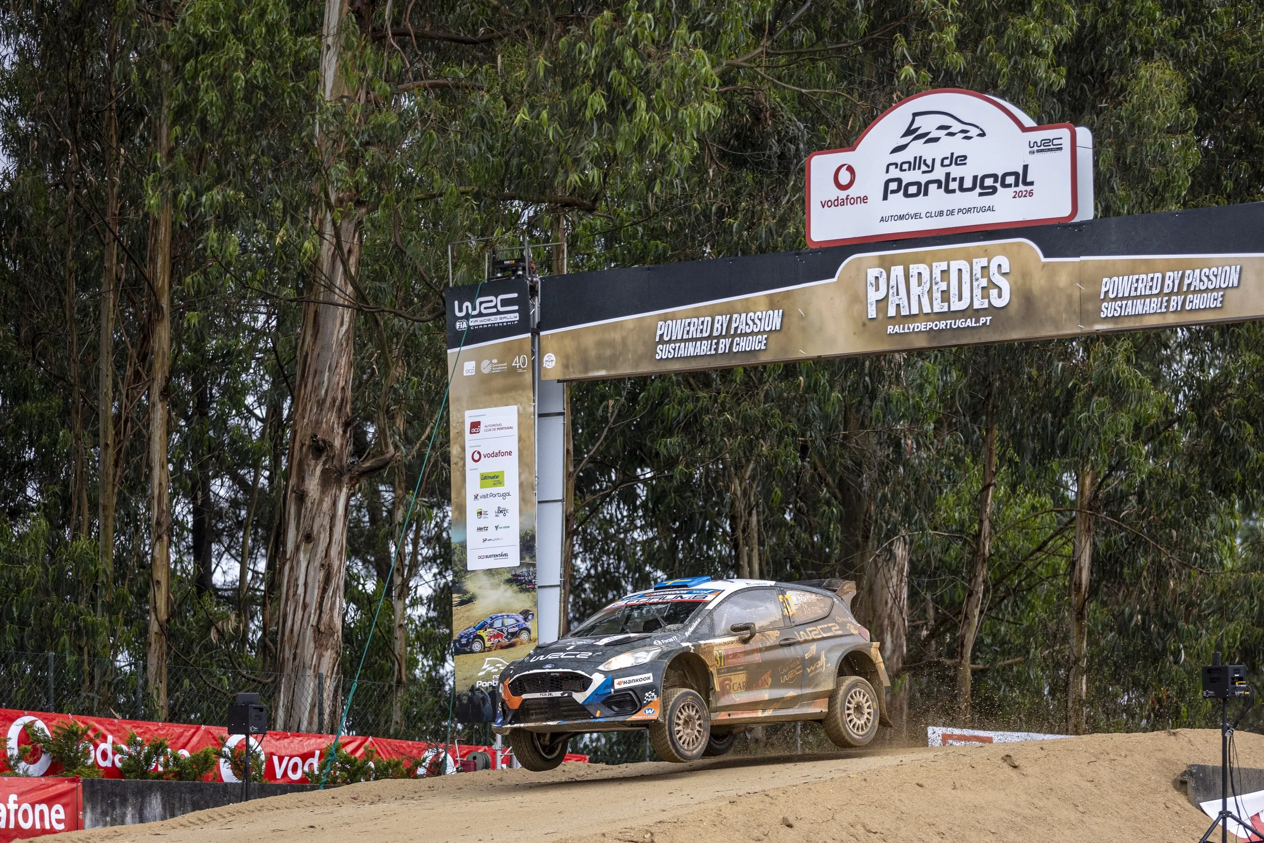 A rally car flying through the air on a dirt track during the Rally de Portugal, with banners and trees in the background.