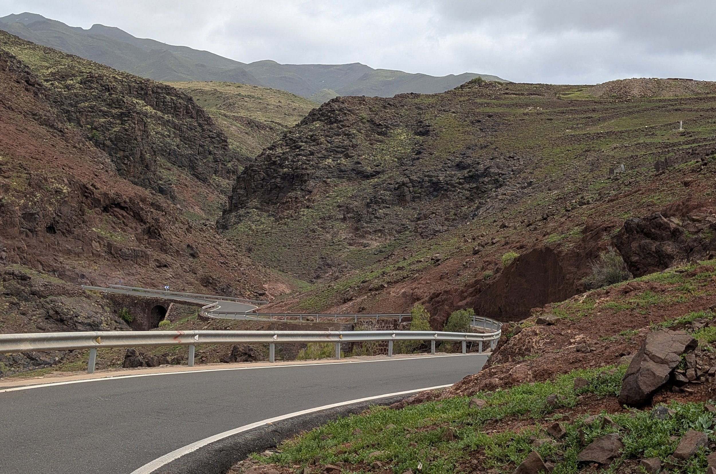 A winding mountain road with guardrails, surrounded by rocky hills and sparse vegetation, under a cloudy sky.