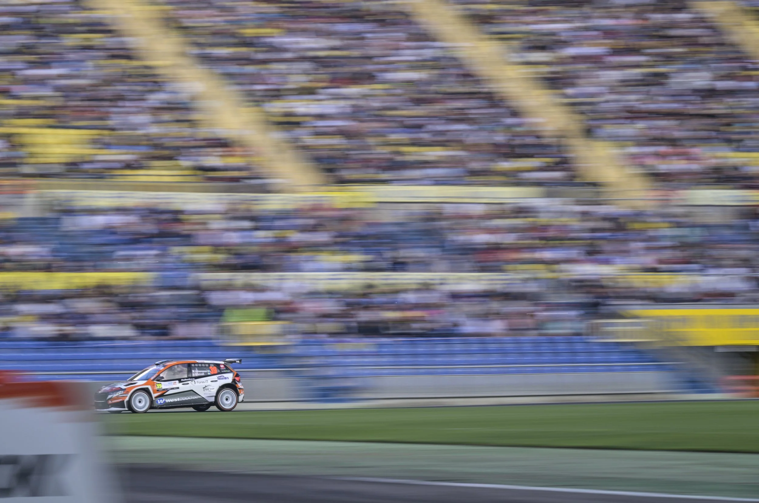 A race car on a track, speeding past blurred spectators in the grandstand. The car is black, white, and orange with various sponsor logos, including one for West 2000cc.