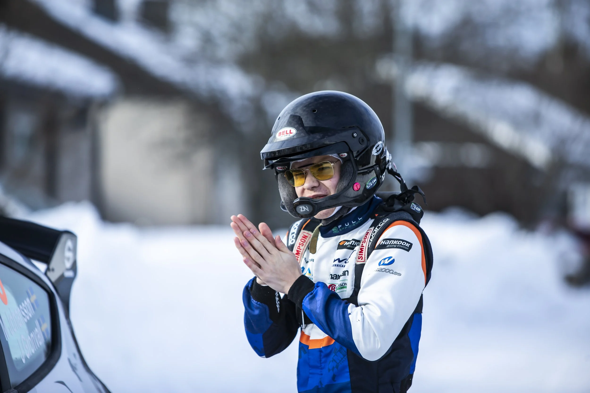 Person wearing a black helmet with yellow-tinted glasses and a racing suit with sponsor logos, standing outdoors in a snowy environment, clapping their hands.