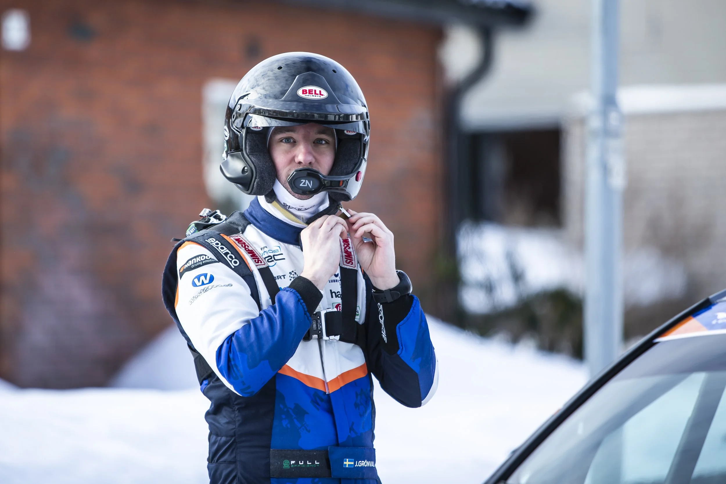 A male race car driver wearing a blue, white, and black racing suit and a black helmet with the Bell logo, standing outdoors in a snowy area near a gray car, adjusting his helmet strap.