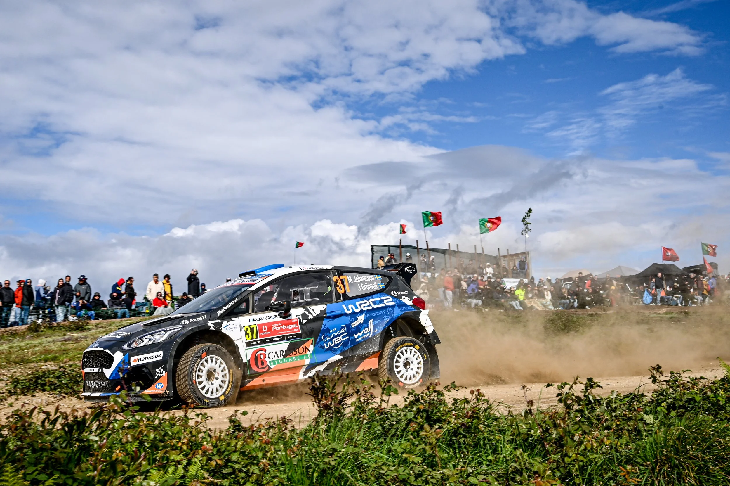 A rally car racing on a dirt track with spectators in the background and flags flying, under a partly cloudy sky.