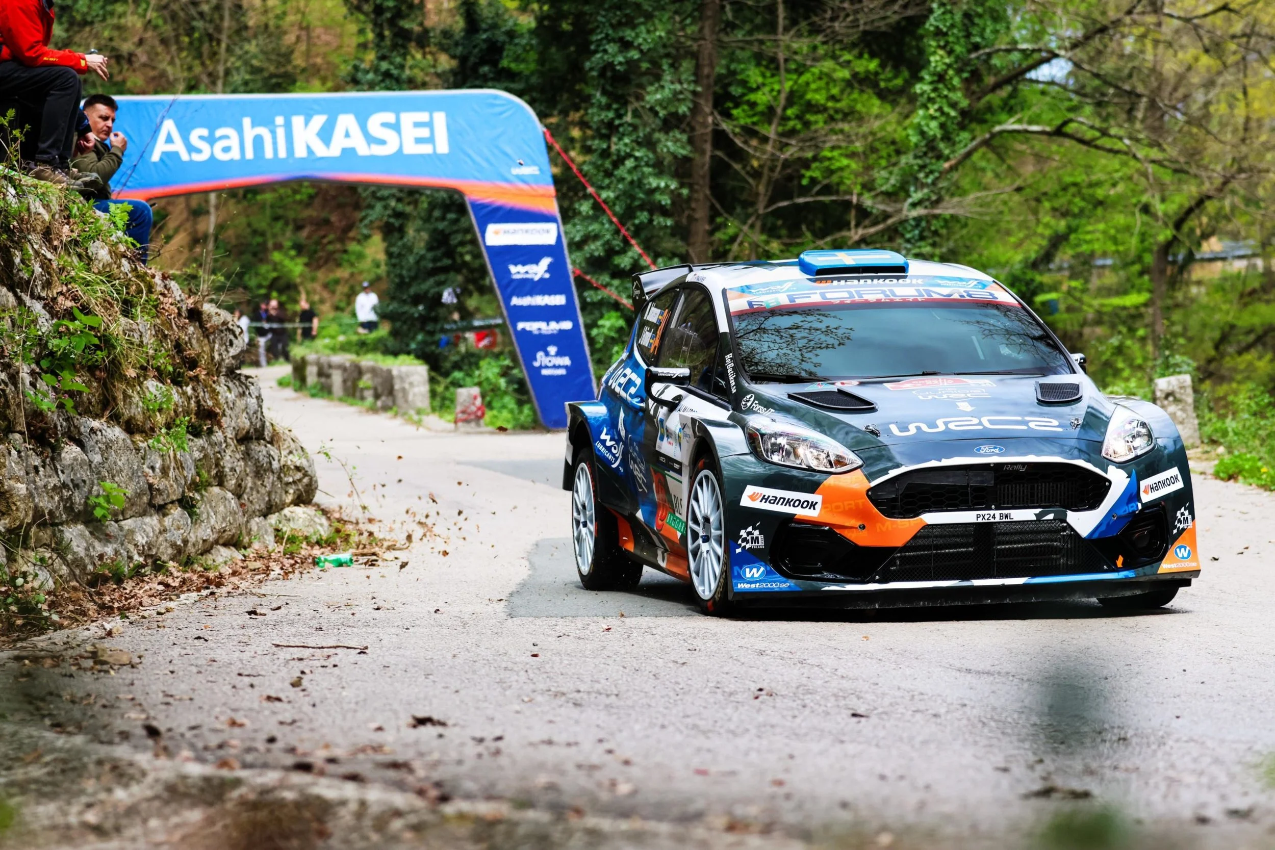 A rally car on a narrow, winding road surrounded by greenery. There are two people sitting at the edge of a rock wall watching the race, and a banner with the text "AsahiKASEI" is displayed across the road.