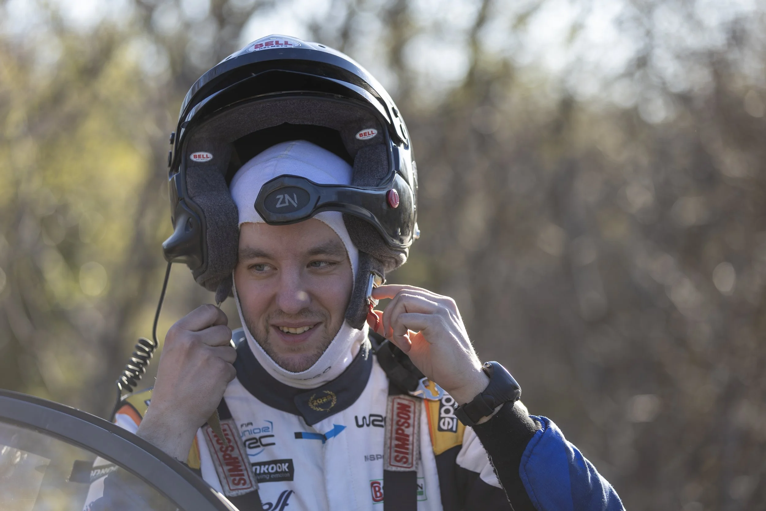 A young race car driver in a racing suit with sponsor patches, adjusting his helmet strap outdoors with a blurred background of trees.