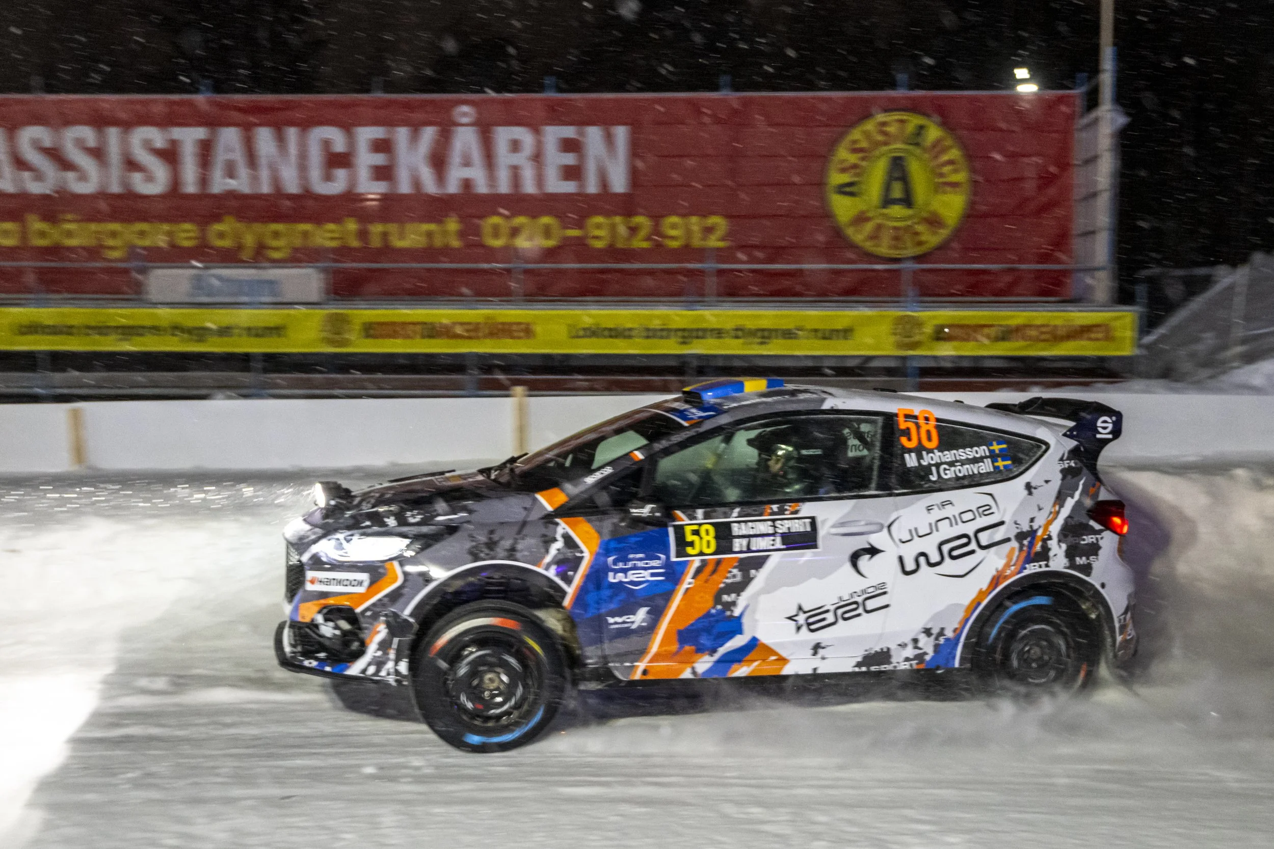 Race car driving on snowy track at night with icy snow falling, advertisements on billboard in background.