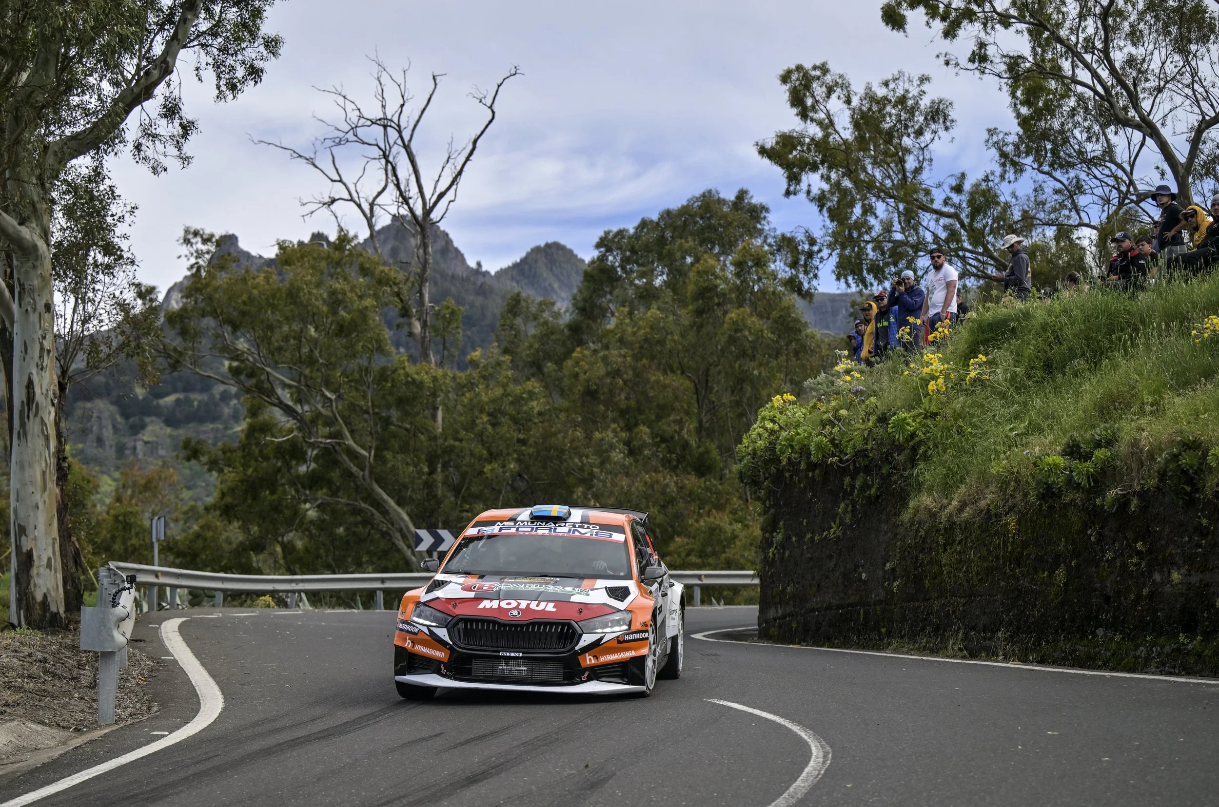 A rally car with an orange, black, and white design racing on a winding road through a forested hilly area with spectators watching from the roadside.