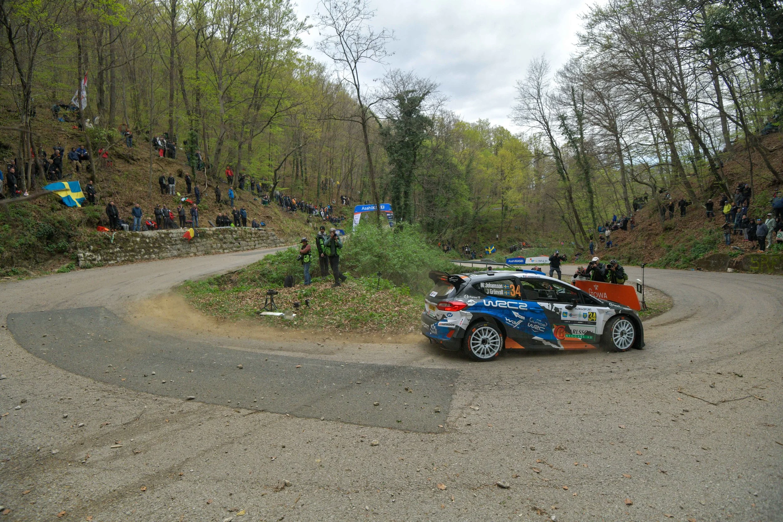 A rally car speeds around a corner on a dirt road, with spectators standing on a hillside and along the roadside behind safety barriers, in a forested area with tall trees and overcast skies.