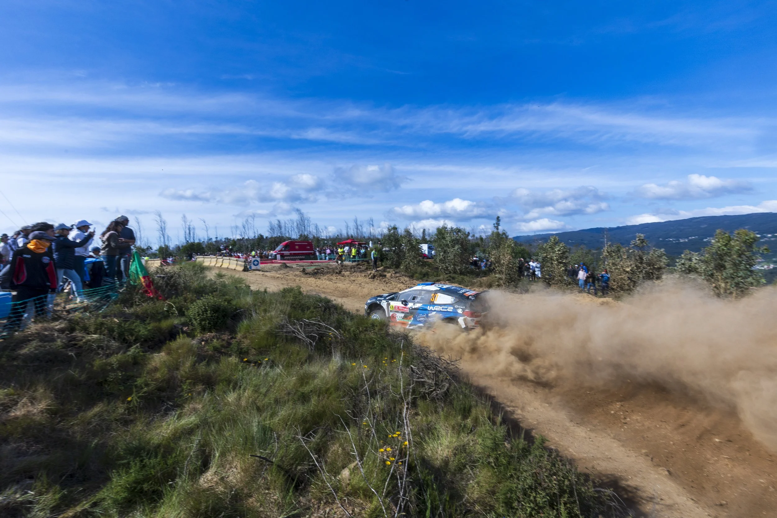 Rally car race on a dirt track with spectators watching, dust and dirt flying, scenic landscape with mountains and blue sky in the background.