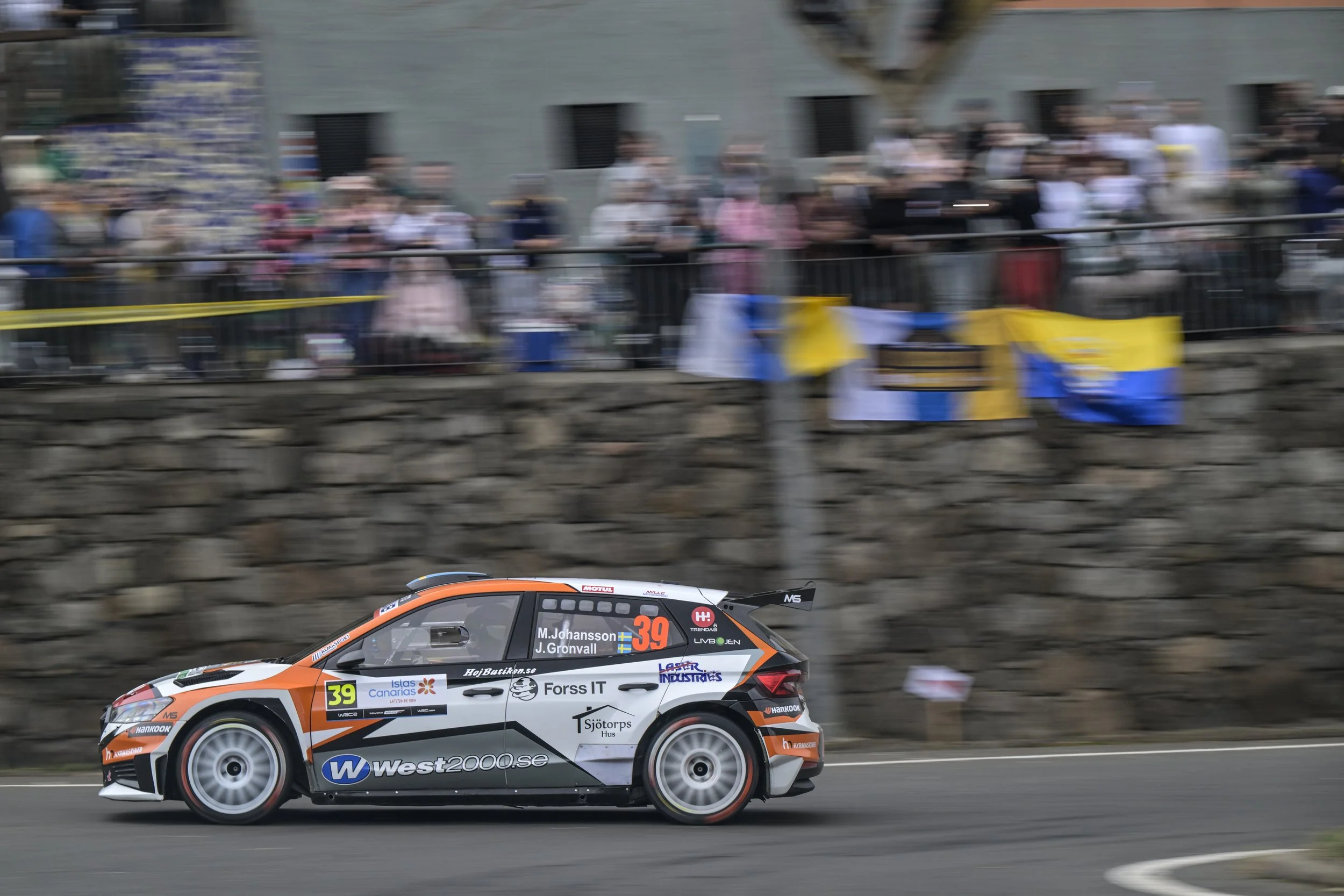 Race car with orange, white, and black design speeding on a track, with a blurred crowd of spectators in the background.