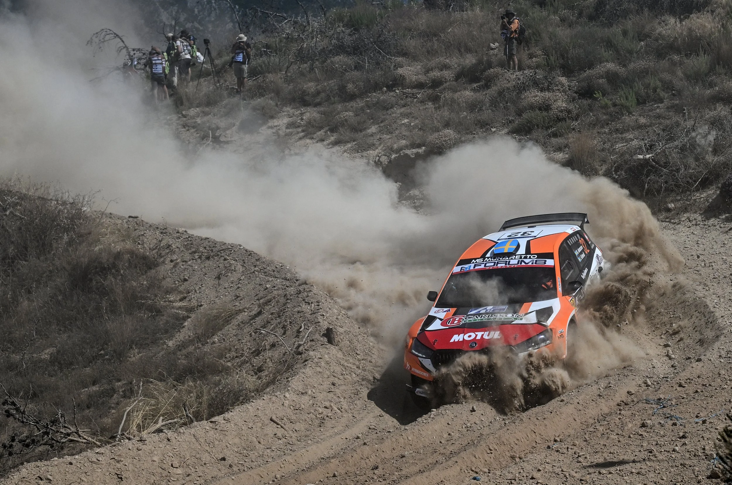 A rally car kicking up dust on a dirt trail in a dry, mountainous area with sparse vegetation. There are several photographers and spectators on the hillside capturing the event.