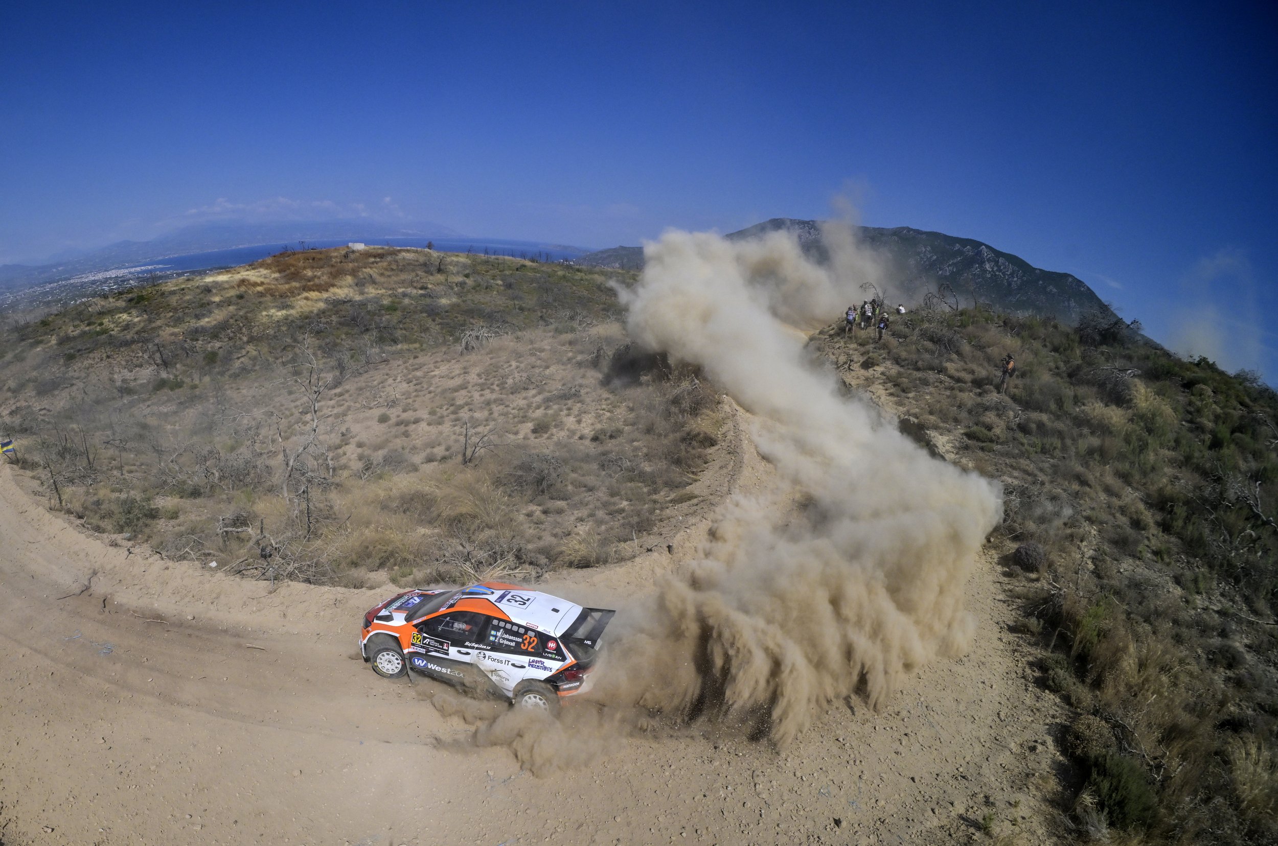 A rally car racing on a dirt trail in a mountainous area, with dust clouds and a few photographers capturing the event.