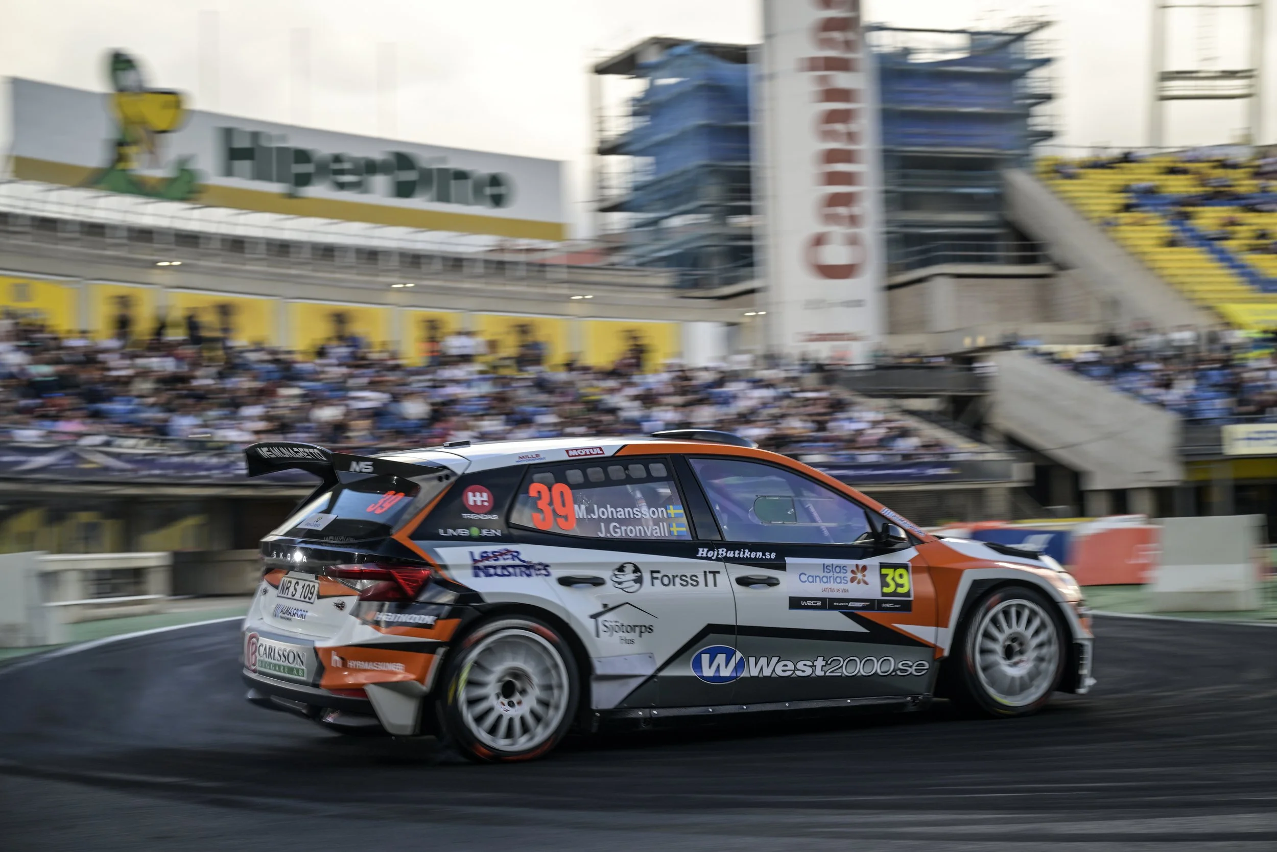A race car with the number 39 and various sponsor logos, speeding on a track during a racing event, with a blurred crowd of spectators and stadium background.