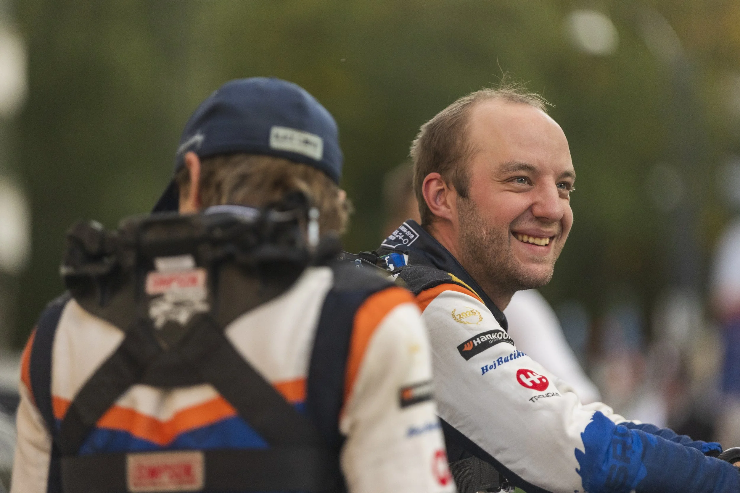 Two race car drivers wearing racing suits and gloves, smiling and engaging with each other, outdoors with blurred greenery in the background.