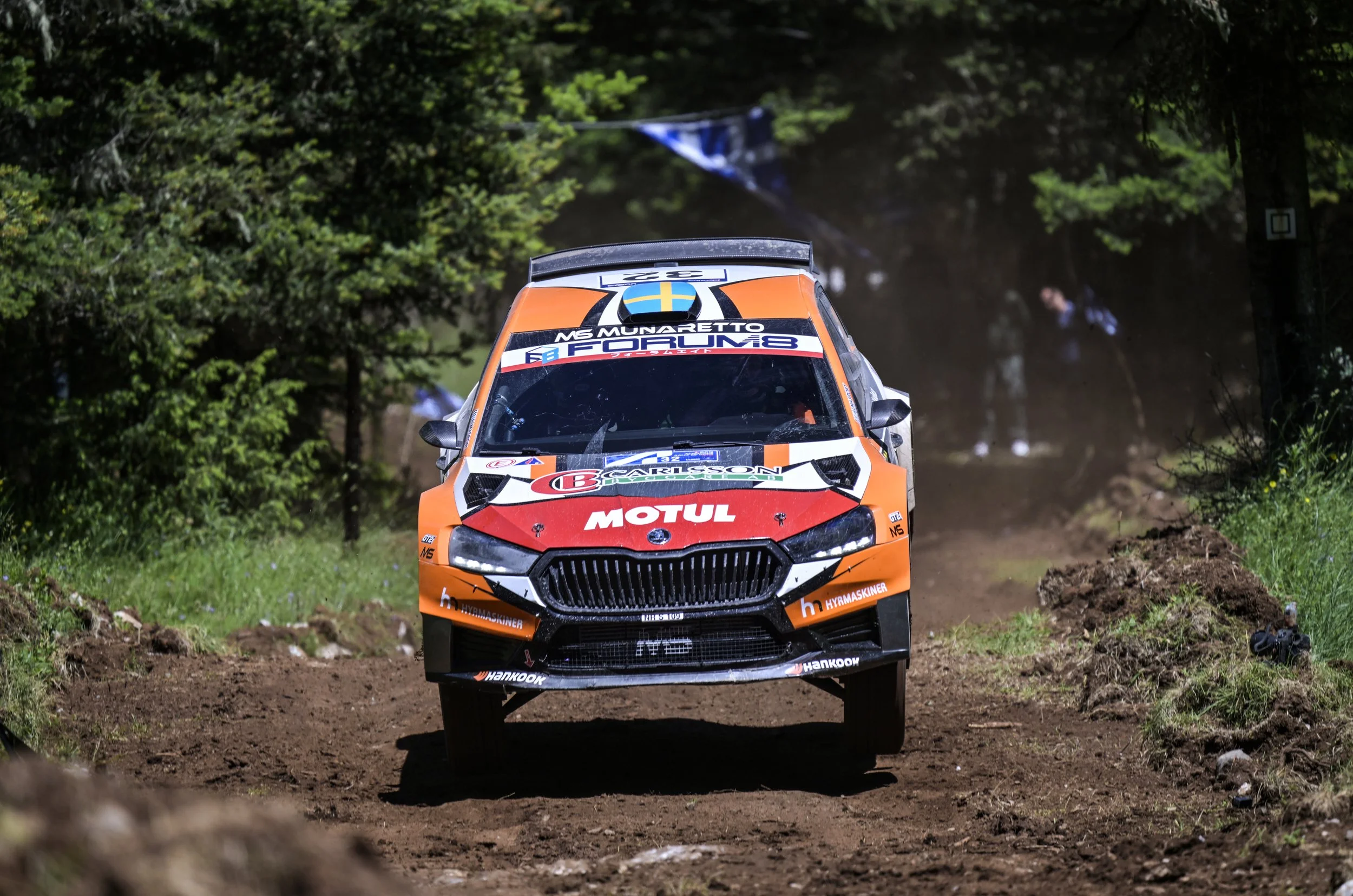 An orange and black rally car driving on a dirt forest trail with trees on both sides and a blue and white flag in the background.