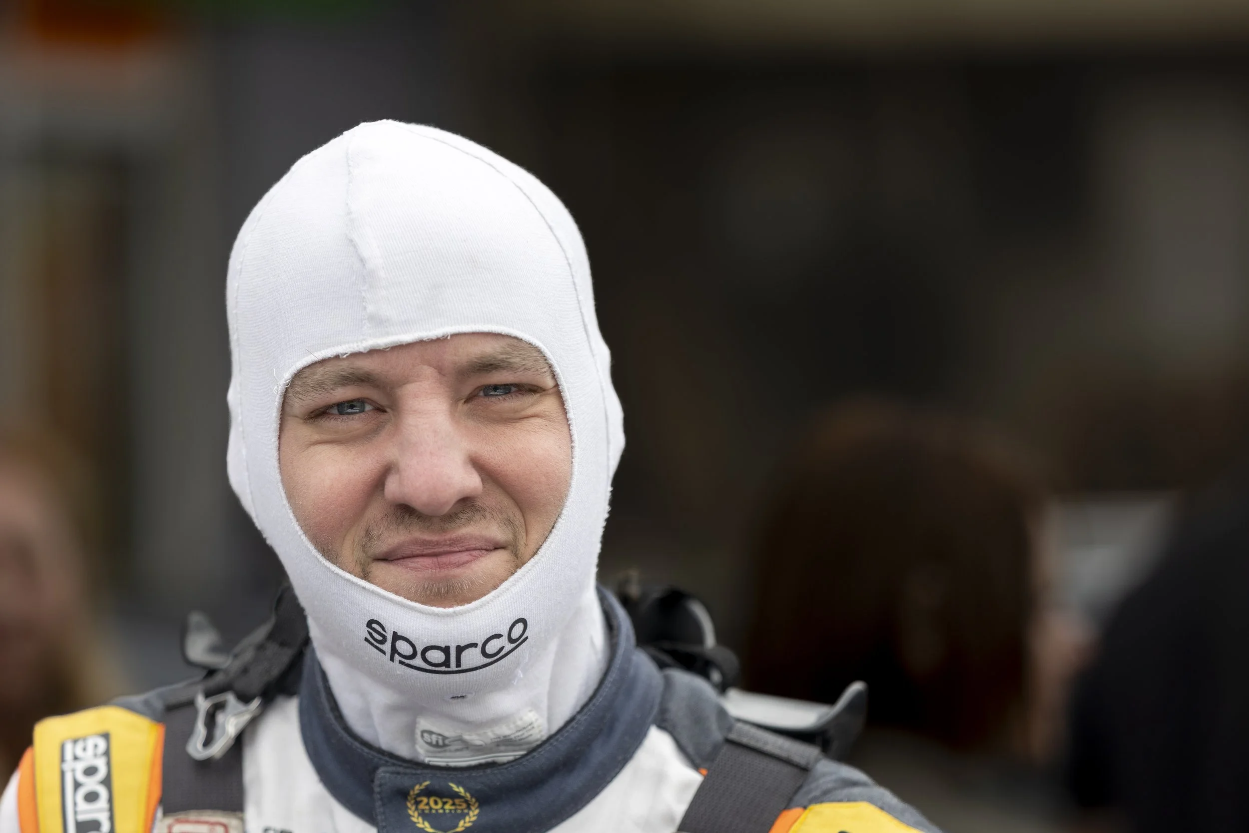 Close-up of a smiling young man in racing gear and a white balaclava, with a dark background.