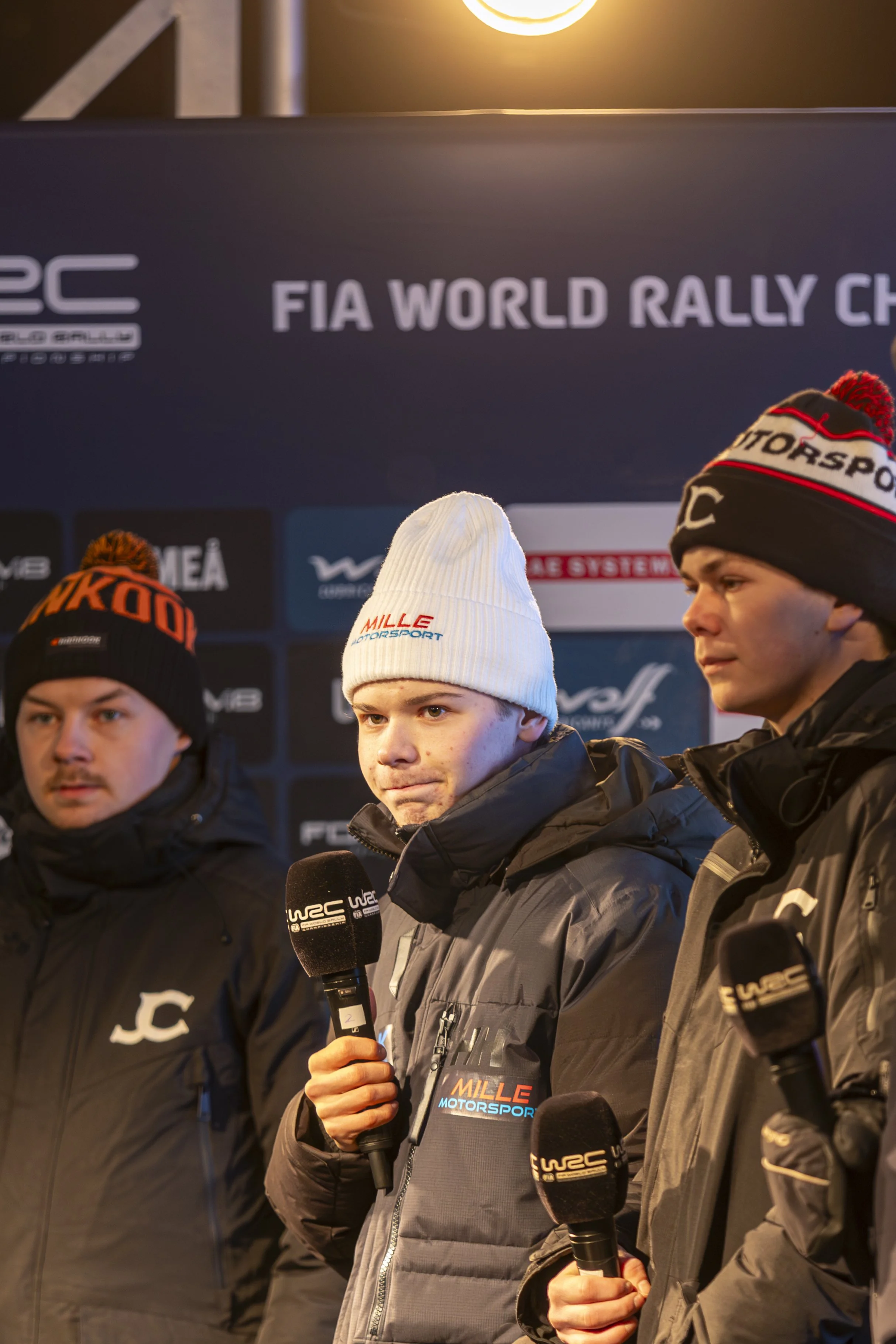 Young male rally drivers being interviewed at a rally event, wearing winter jackets and beanies, with FIA World Rally Championship branding visible in the background.