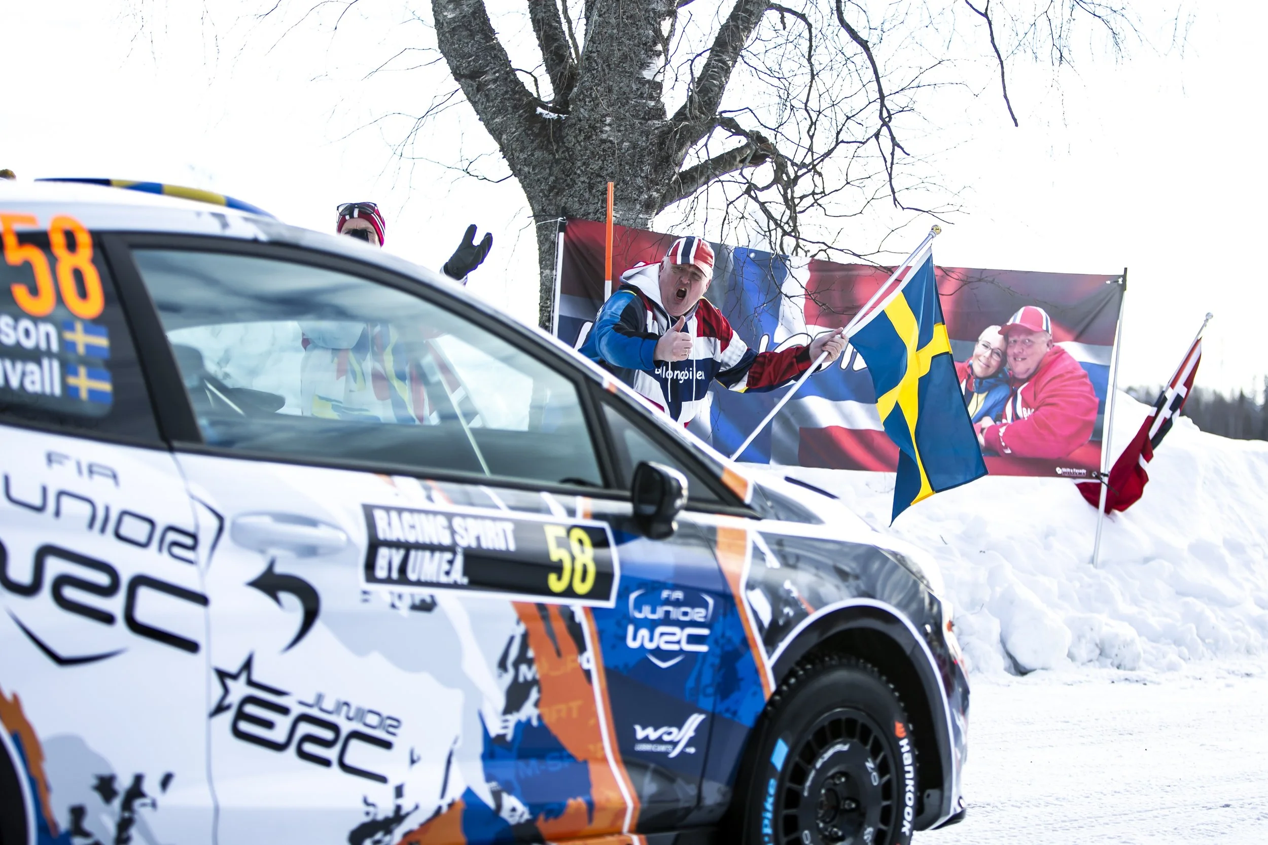 Race car with sponsorship logos and number 58, on snow-covered ground, with a person in rally attire standing behind it, waving and giving a thumbs-up, alongside flags and a large banner in the background.