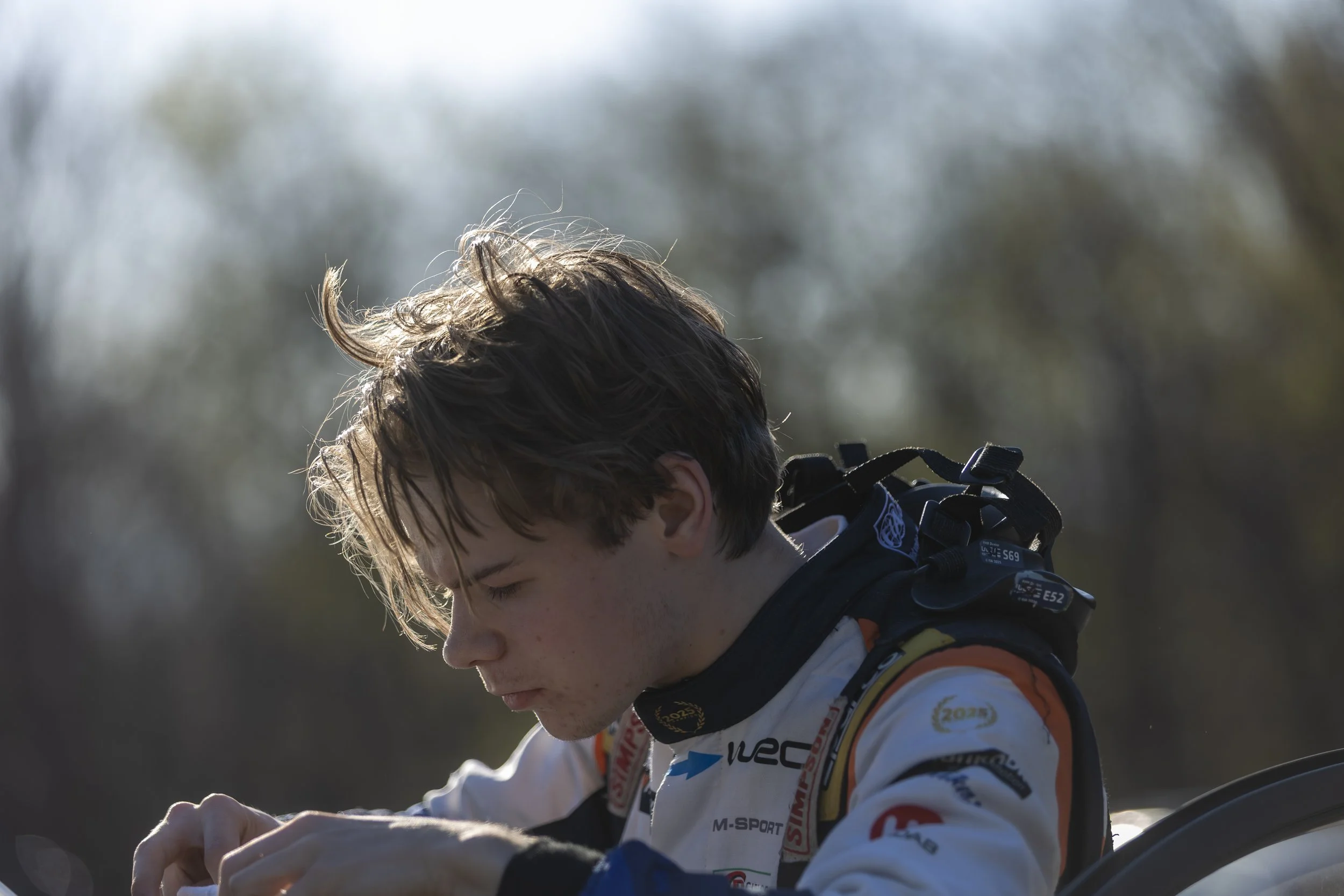 Close-up of a young male racing driver with brown hair, wearing a white racing suit with sponsor patches, outside on a sunny day.