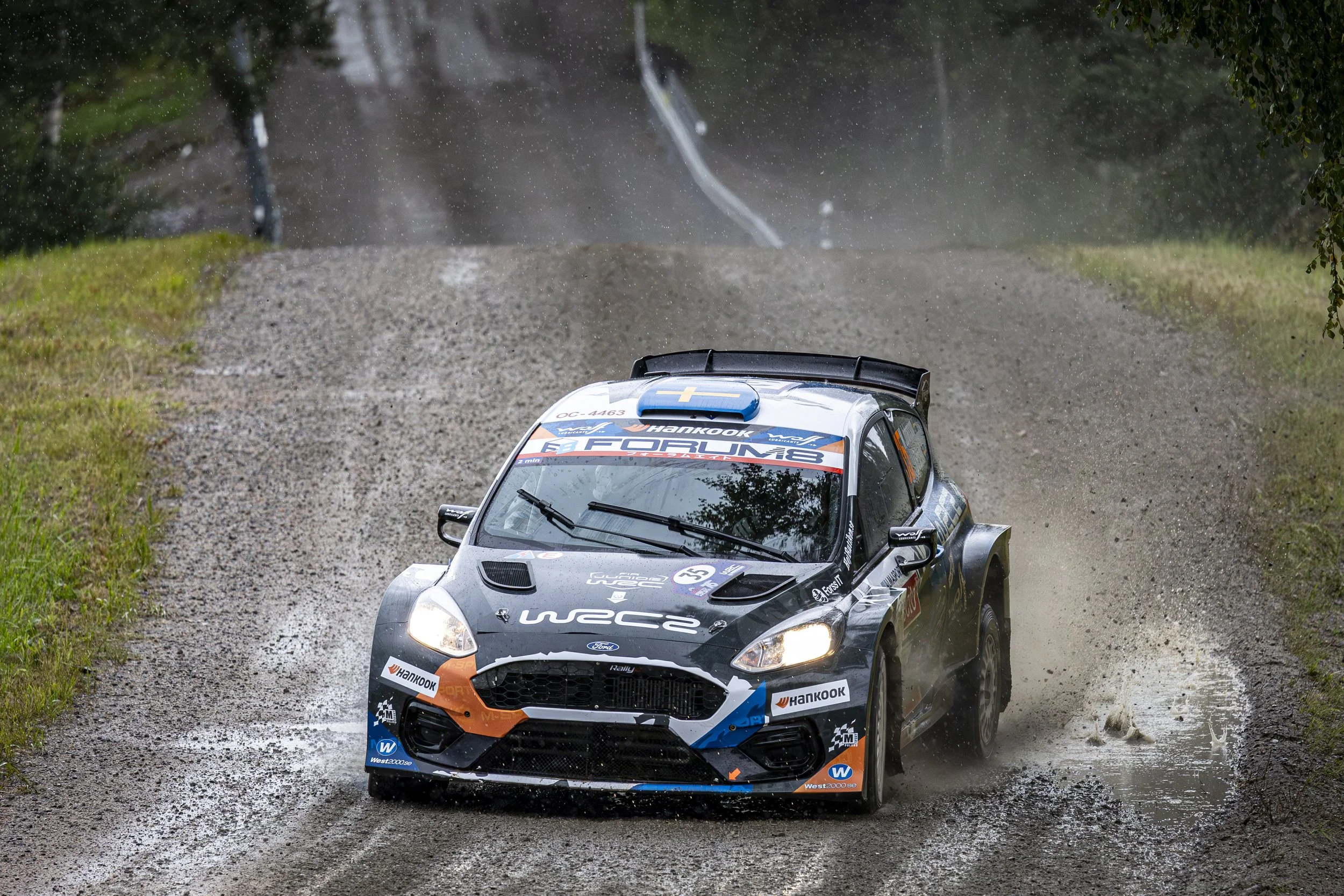 A rally car racing on a muddy dirt road during a race, with water splashing around and lush green trees in the background.