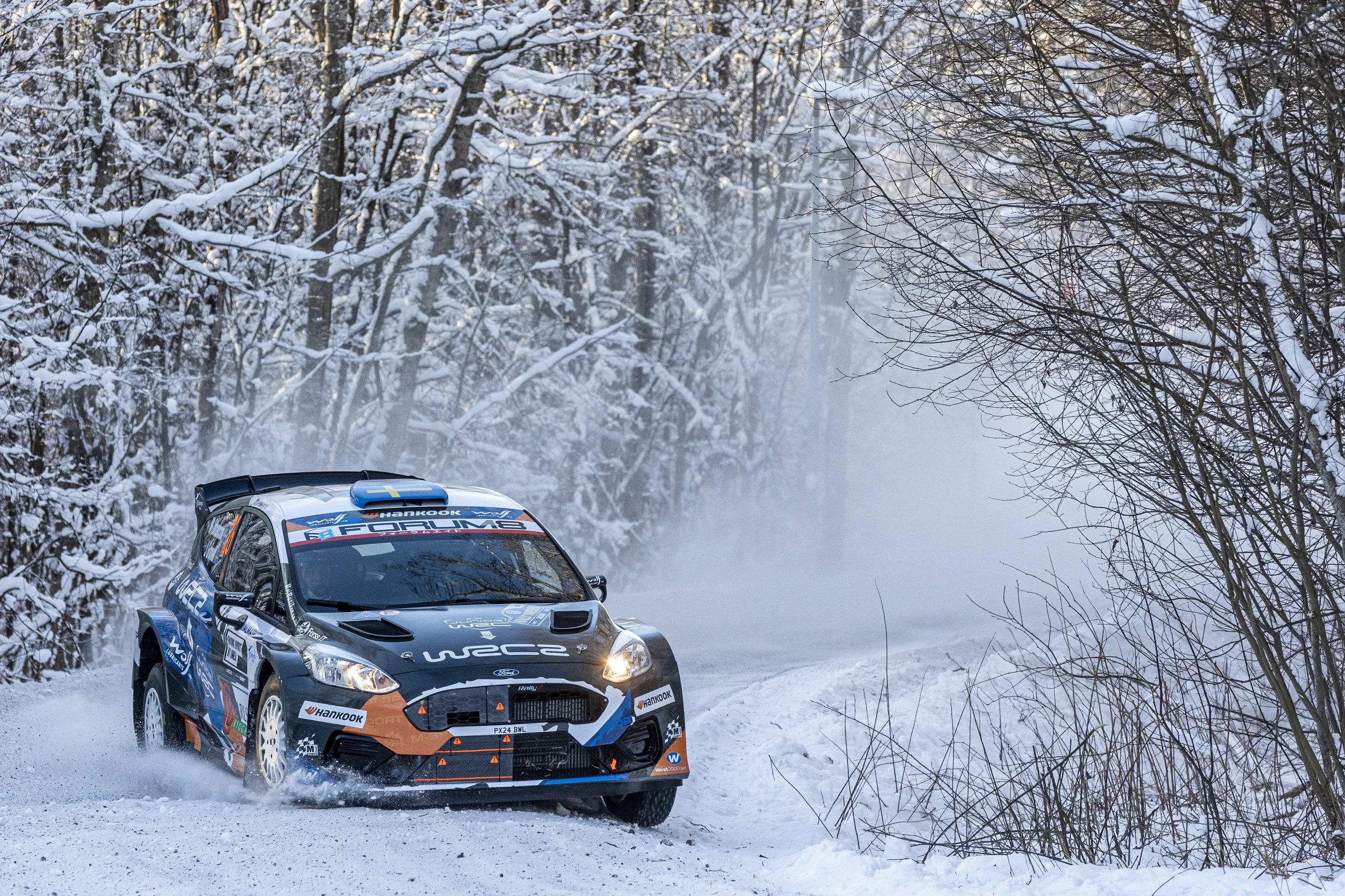 A rally car speeding on a snow-covered road surrounded by snow-covered trees, with visible tire tracks and snow flying off the tires.