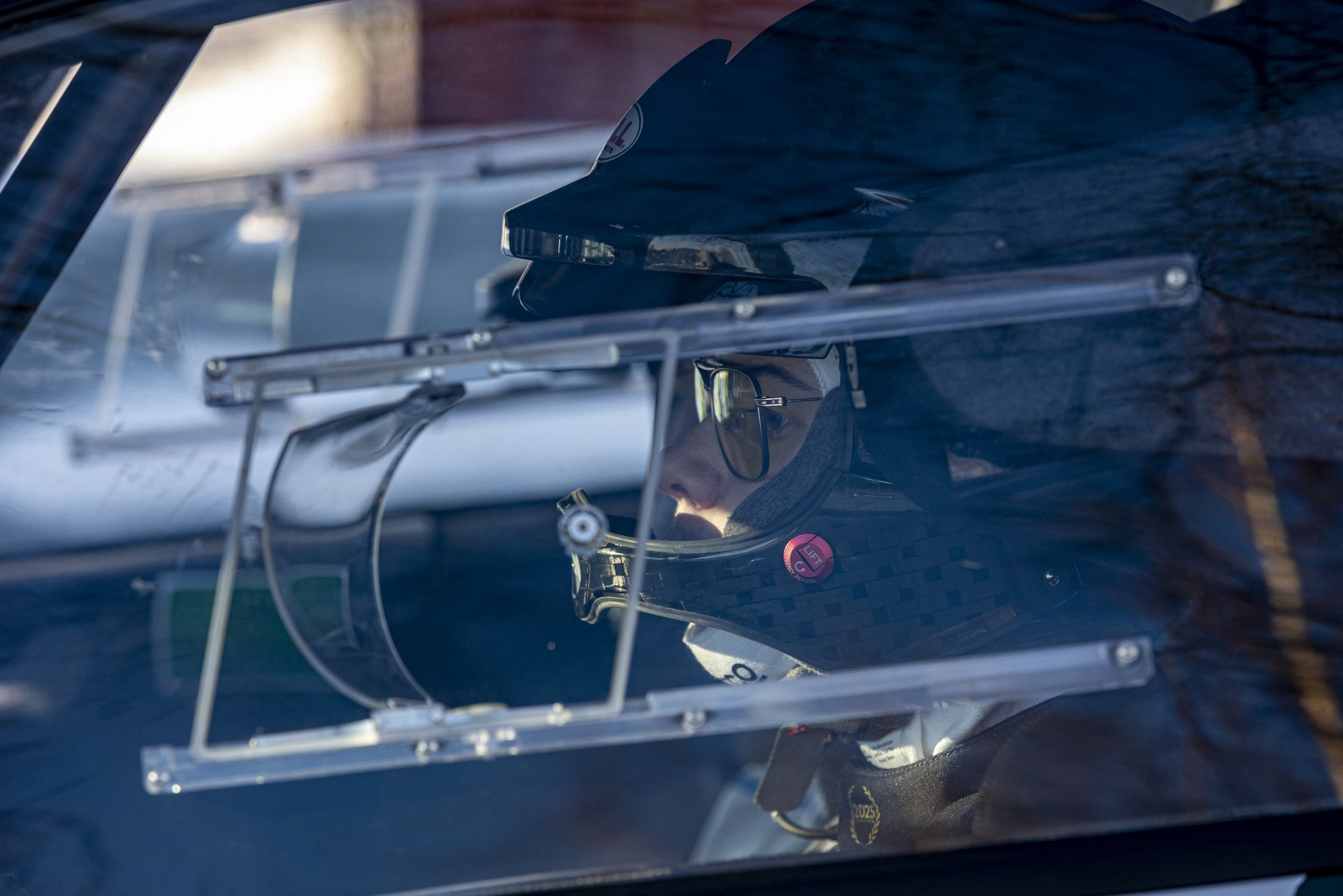 A race car driver wearing a helmet and safety gear inside a race car, with the car's window reflecting the outside environment.