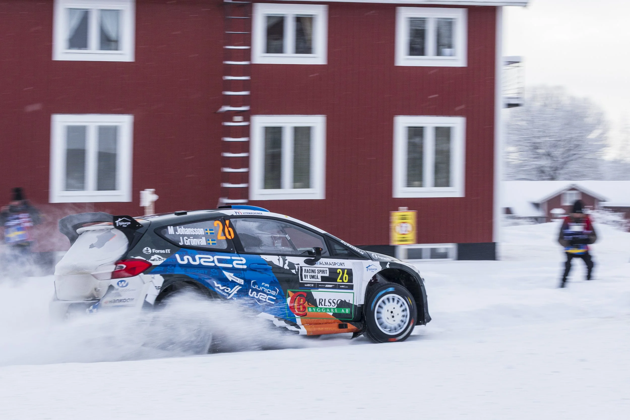 A rally car speeding on a snowy road, kicking up snow behind it, with a red building and snowy trees in the background.