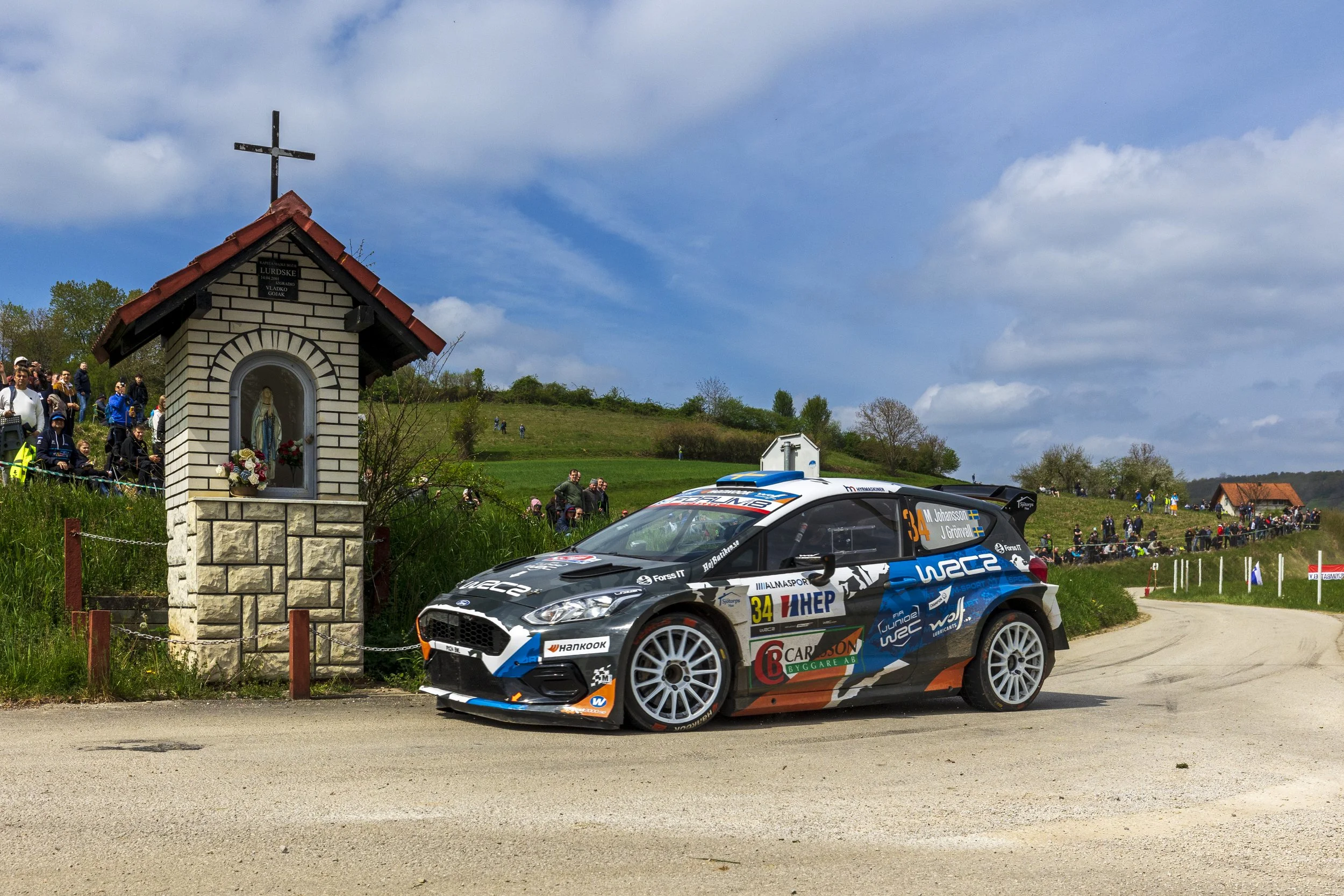 A rally car with a black, blue, and orange design speeds past a small shrine with a statue of the Virgin Mary, while a crowd of spectators watch from a grassy hill under a partly cloudy sky.