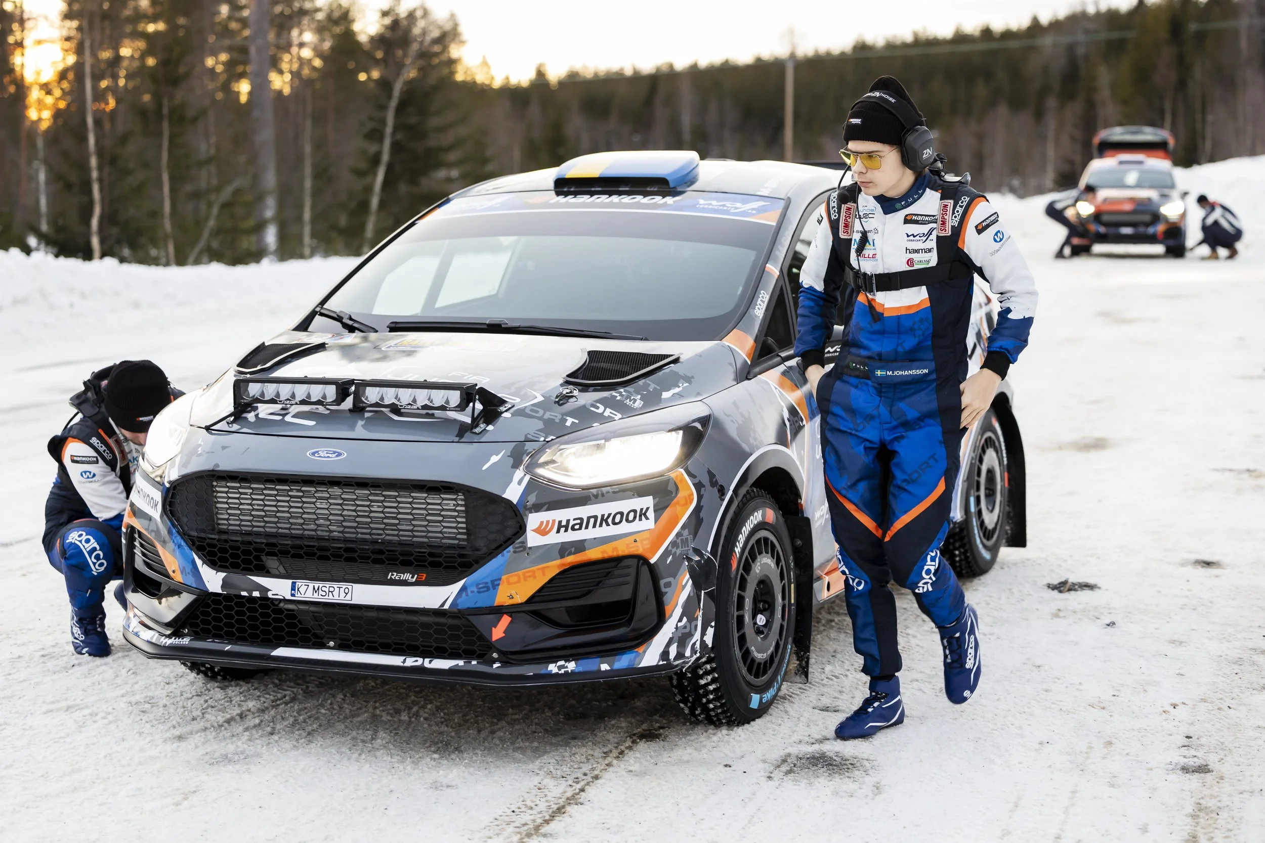 Three rally race car drivers in racing suits standing and kneeling next to a black and orange rally car on a snowy road with trees in the background at sunset.
