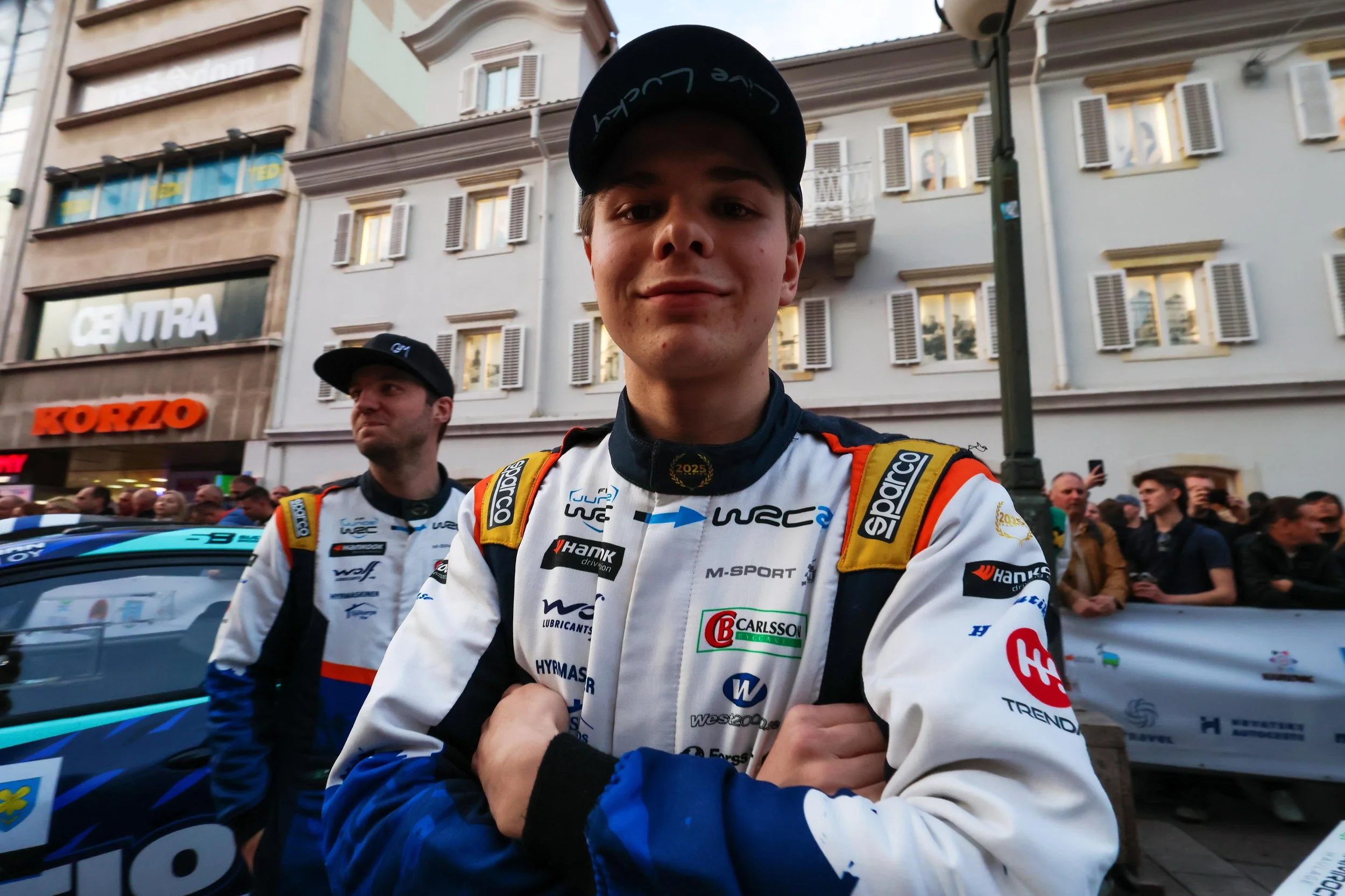 Young male race car driver in a racing suit taking a selfie in a city street with spectators and buildings in the background.