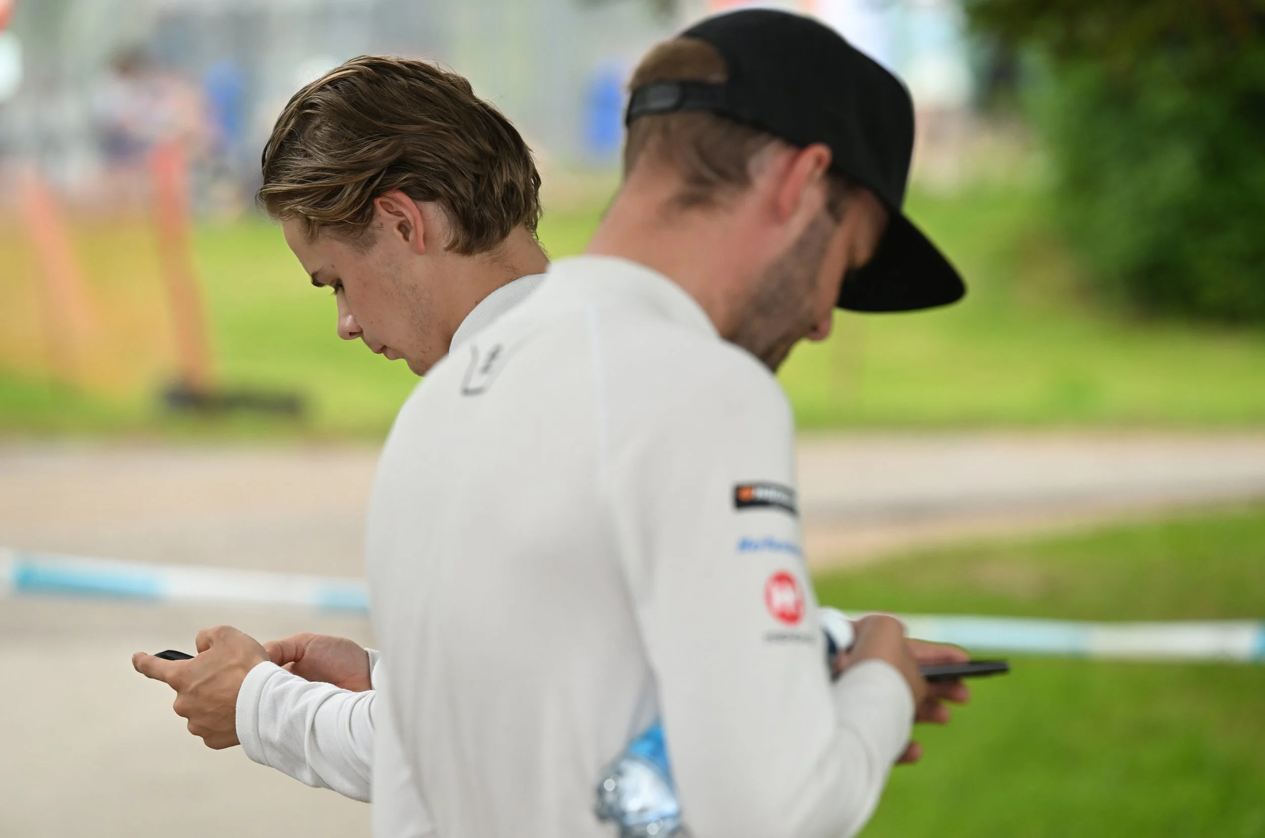 Two young men standing outdoors, both looking at their smartphones and dressed in white sports jackets, with one wearing a black cap. Blurred background with greenery and other people.