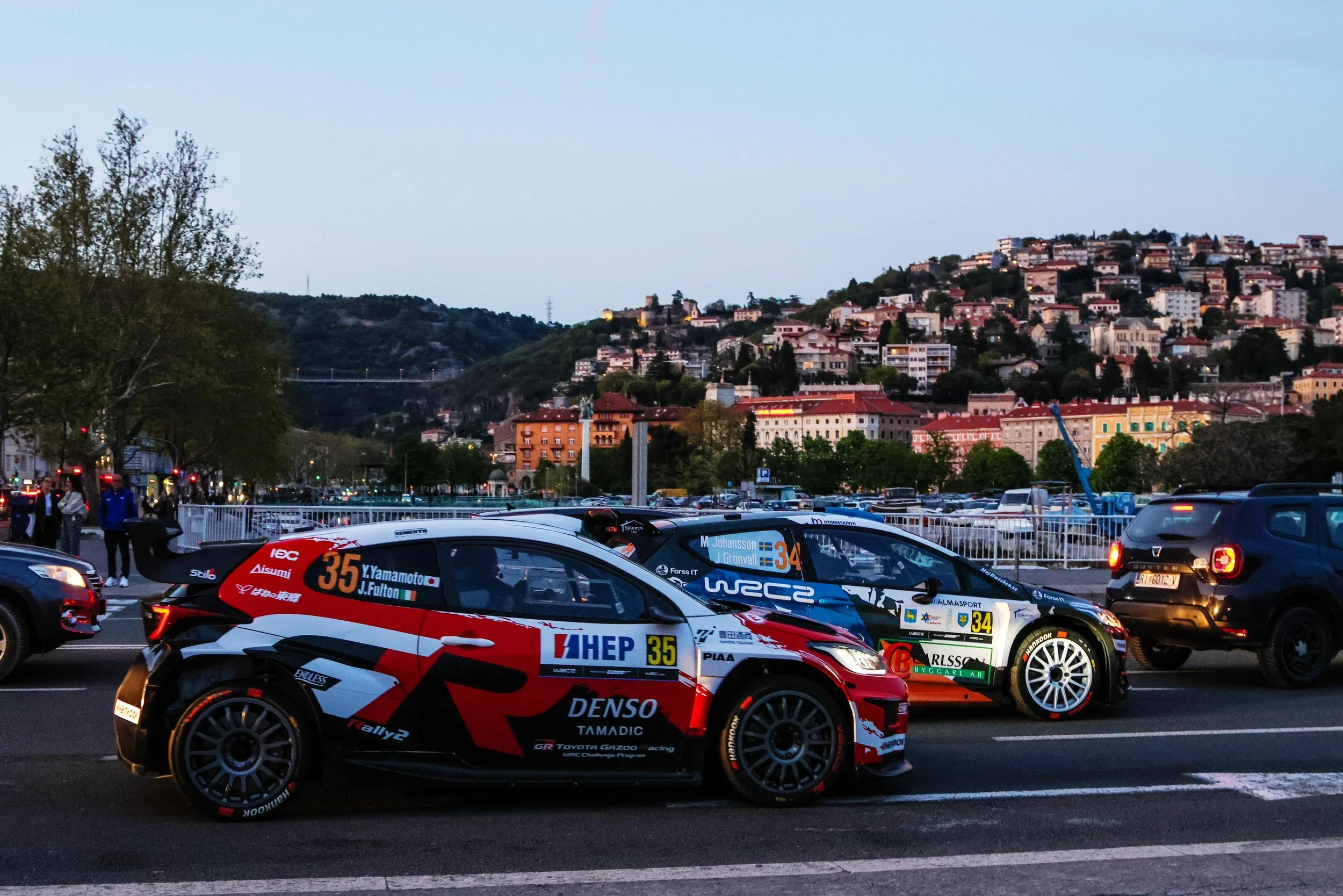 Rally cars parked in a lot with a city hillside in the background at dusk.