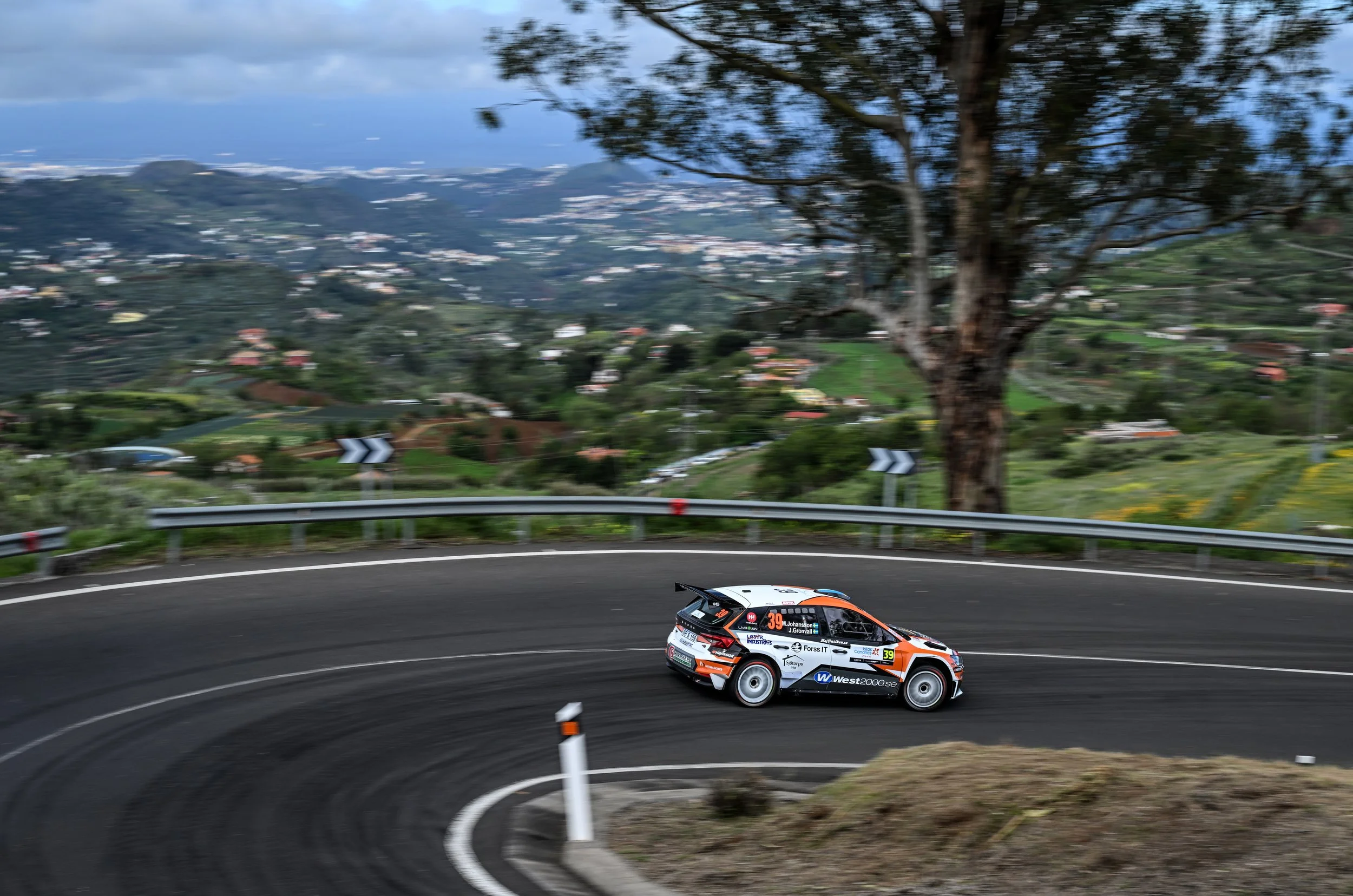 A rally car racing on a winding mountain road with a scenic landscape of trees, hills, and clouds in the background.
