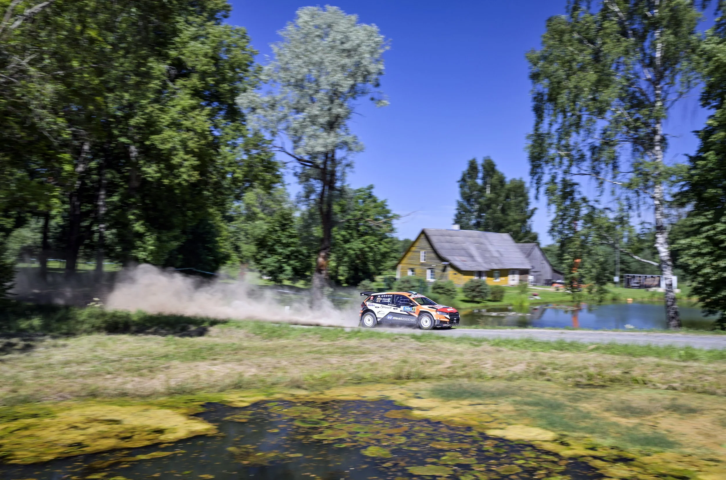 A rally car speeding on a dirt road beside a pond, with dust trailing behind it, surrounded by green trees and a yellow house in the background under a clear blue sky.