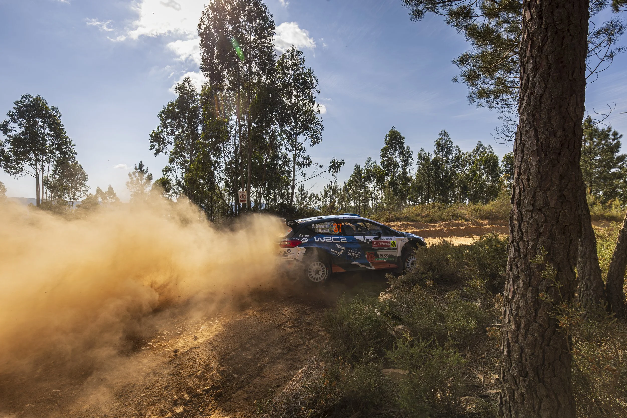 A rally car speeding on a dirt road in a forest, kicking up dust with tall trees and a bright blue sky with some clouds overhead.