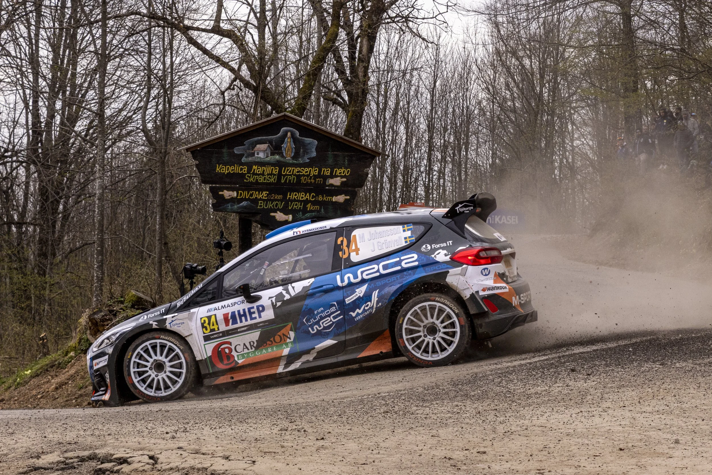 Rally car speeding on a dirt road surrounded by leafless trees. The car has various sponsor logos and the number 34 displayed.