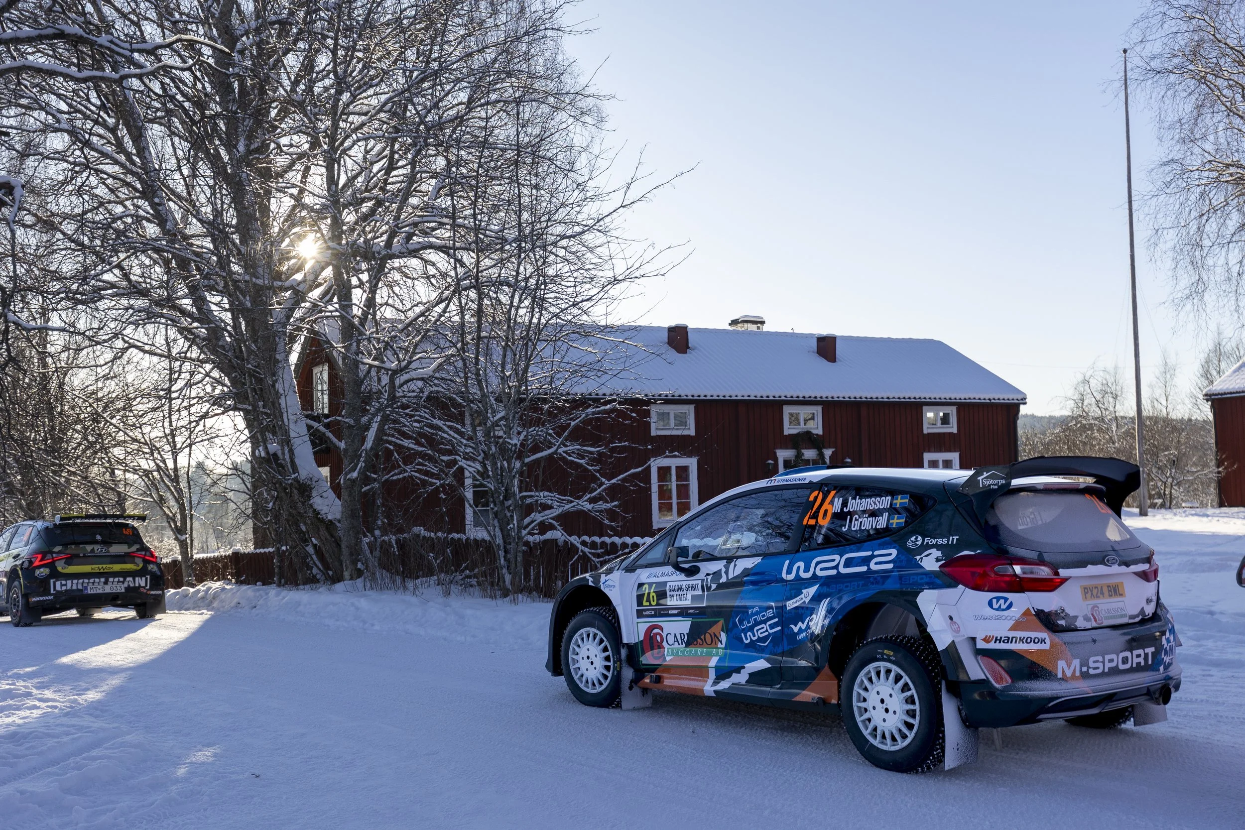 Race cars parked on snow-covered ground near a red house with snow on its roof, leafless trees, and a clear blue sky in a winter setting.